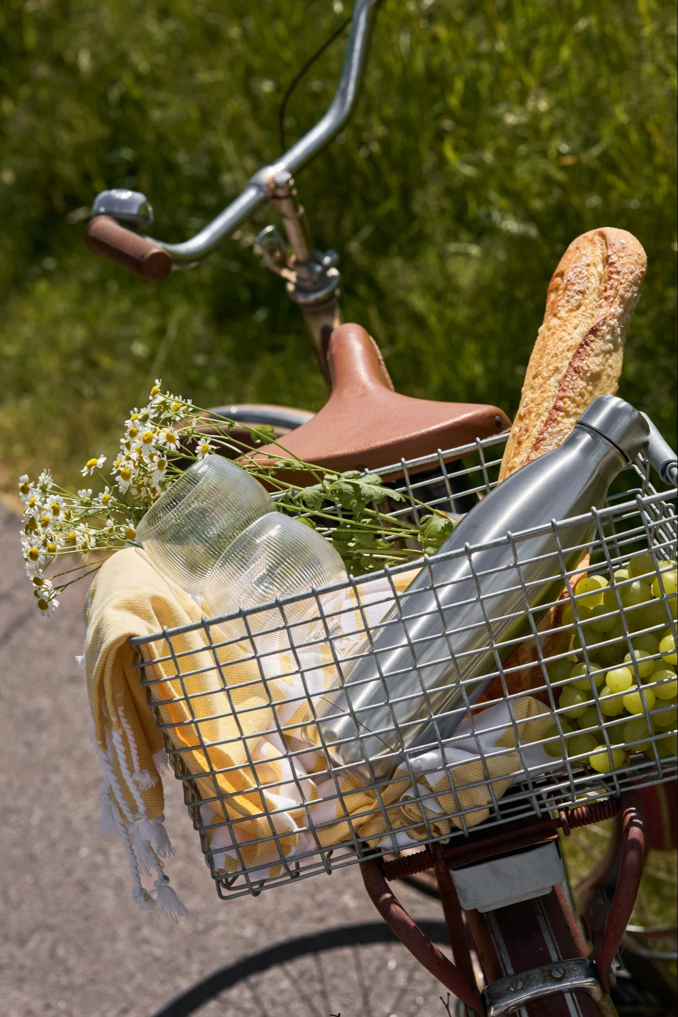 Cykel med en frontkurv fyldt med picnic-ting: brød, grønne druer, en sølvfarvet vandflaske, kopper, et gult håndklæde og blomster.