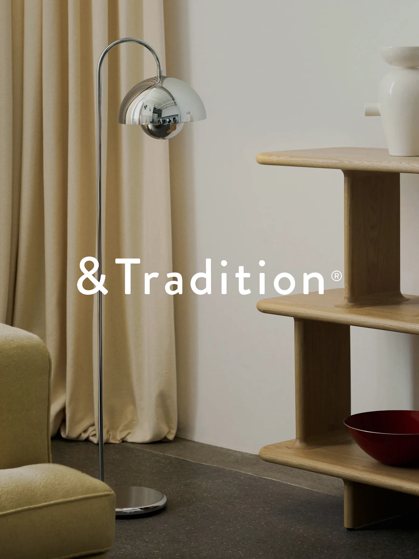 Et moderne interiør med en kromgulvlampe, en etageret hyldereol i lyst træ og en lænestol. &Tradition® logo.