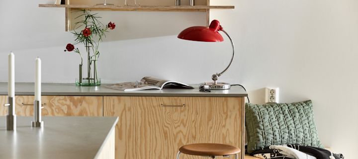 Her kan du se Kaiser bordlampen i rubinrød sammen med Ikeru vasen i børstet stål, begge fra Fritz Hansen, i et moderne krydsfinerkøkken.