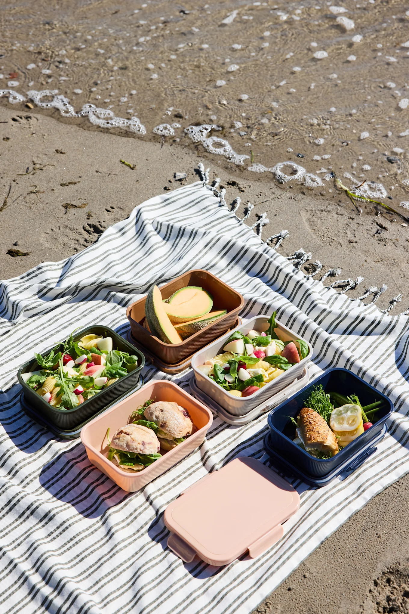 Fem åbne, farverige madkasser med salater, sandwiches og melon, plus en lukket lyserød kasse, på et stribet tæppe på stranden.