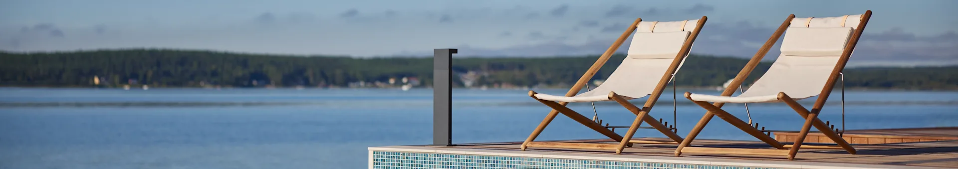 To hvide stof- og træsolstole på en terrasse med udsigt over en sø med skov i det fjerne.