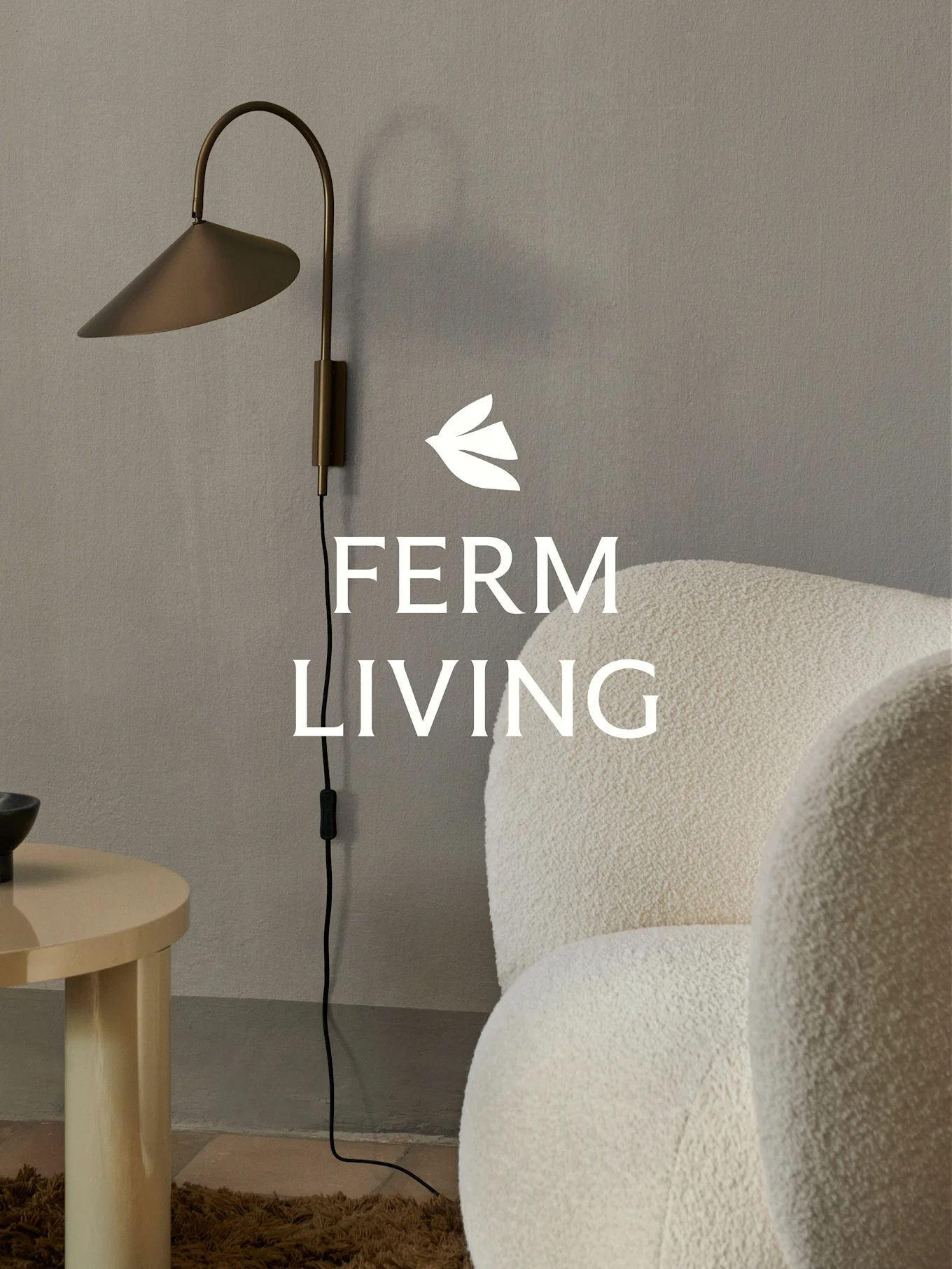 Ferm Living logo over en messing væglampe, en creme bouclé lænestol og et lyst træ sidebord i minimalistiske omgivelser.