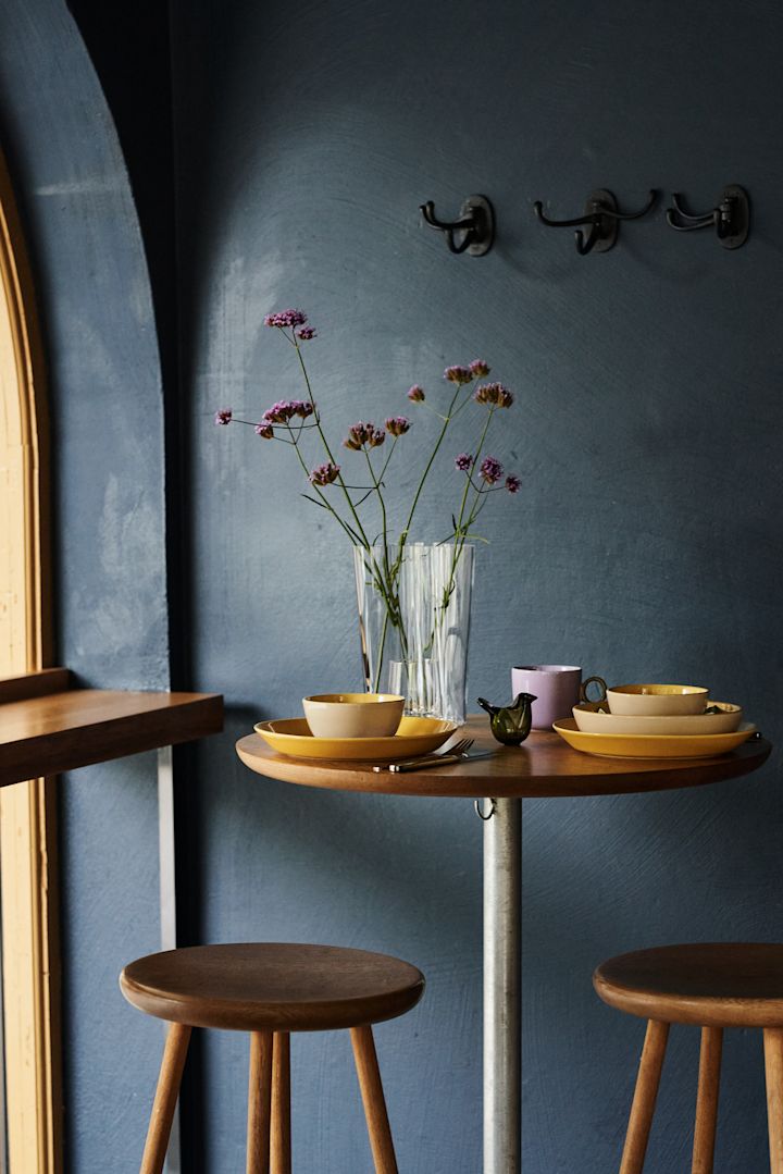 Her kan du se en borddækningside med et retro diner-tema, i gult og lilla i et mørkeblå rum med en klar vase og matchende lilla blomster.