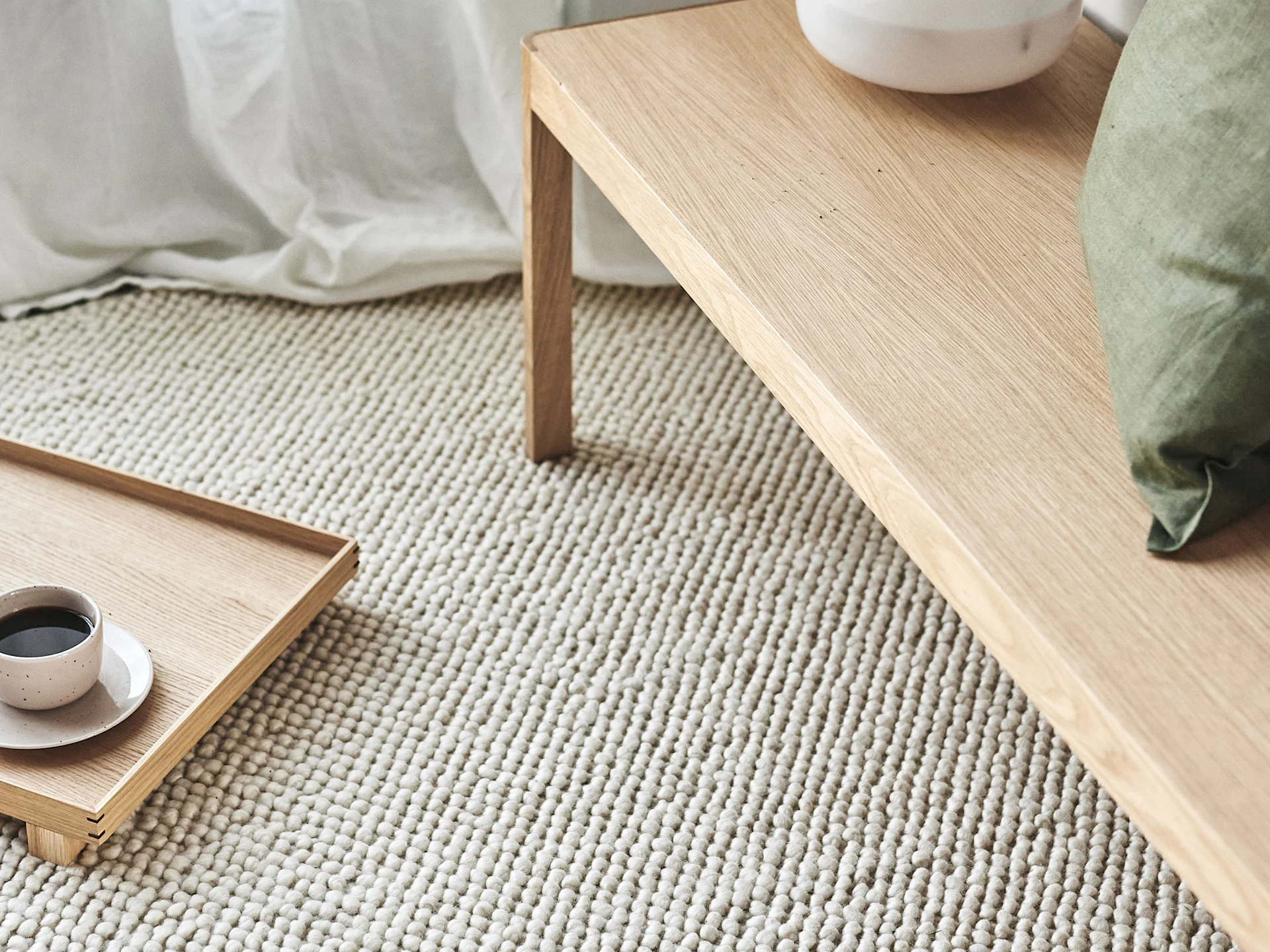 Et lyst sofabord og en bakke med en kaffekop, på et cremefarvet tekstureret tæppe, med en grøn pude.