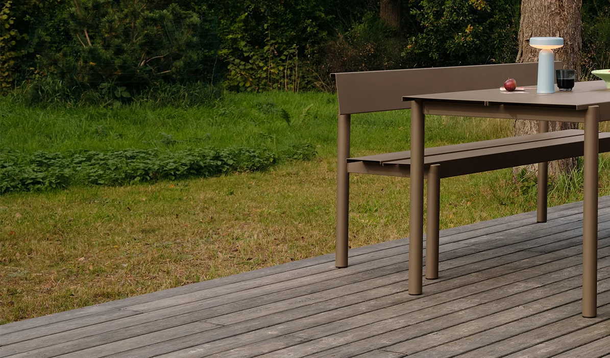 Moderne udendørs spisebord og bænk i taupe på træterrasse, med bærbar lampe og æbler på bordet.