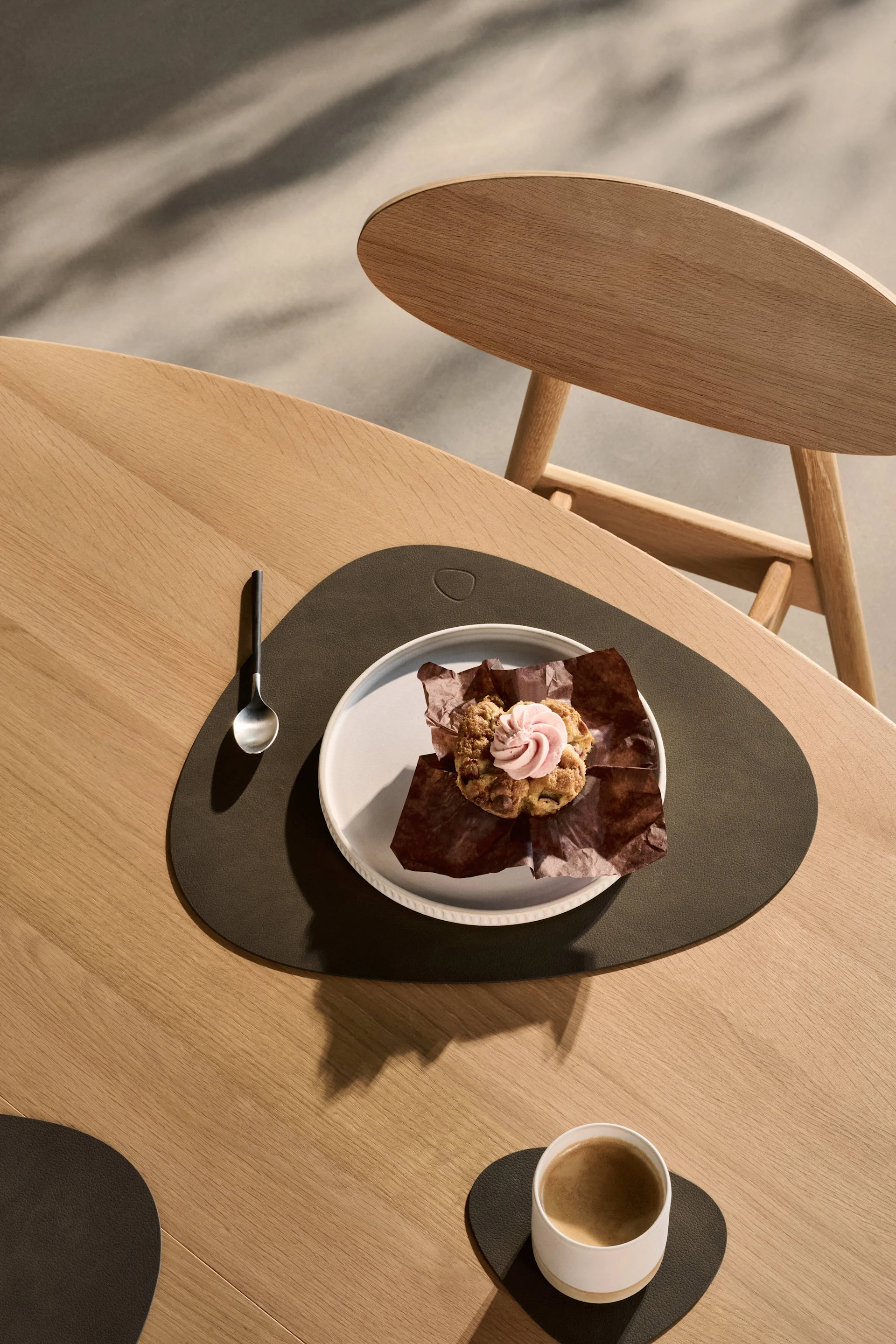 Muffin med lyserød glasur og kaffe på træbord med mørke dækkeservietter og underlag.