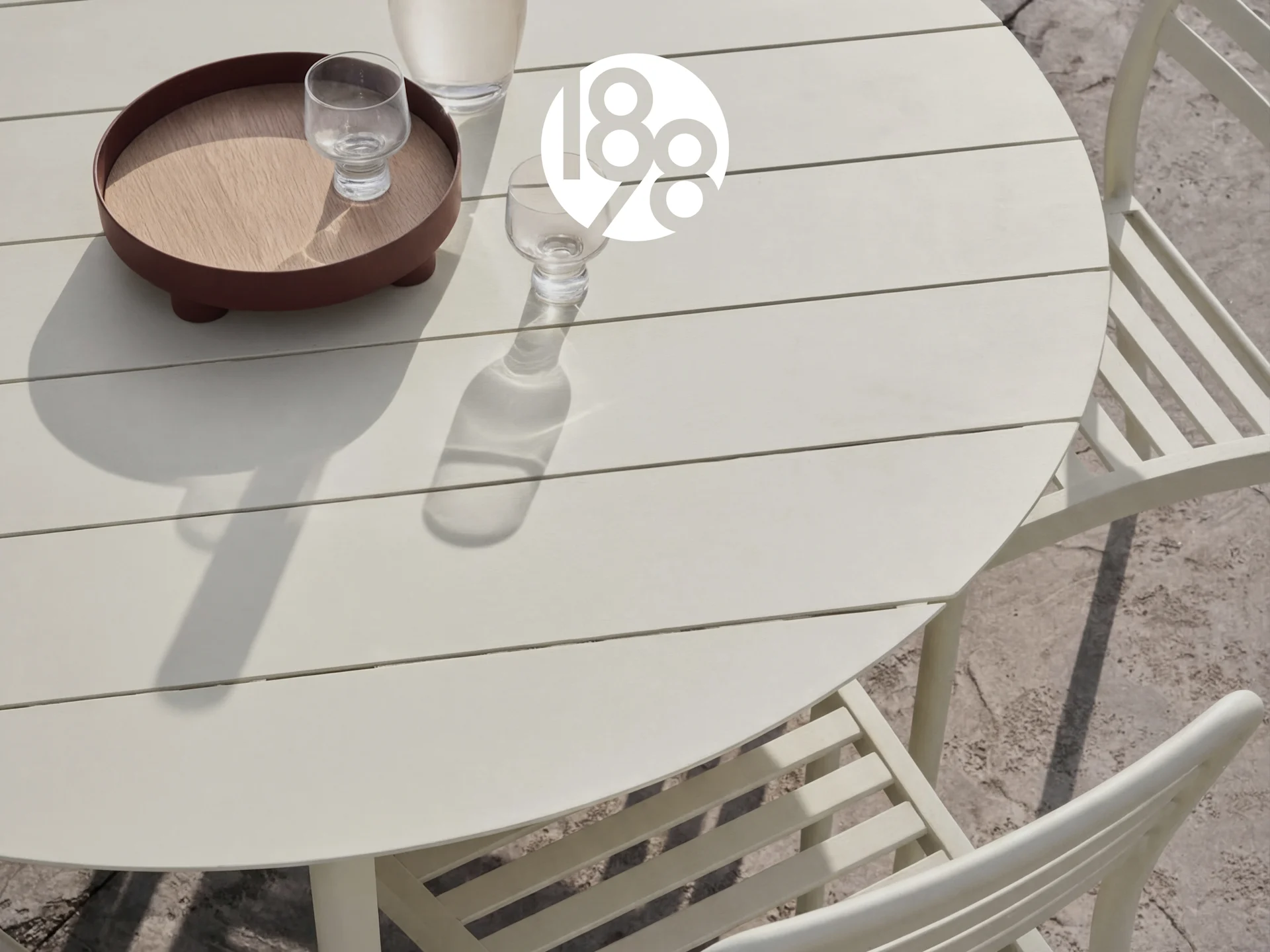 Udendørs rundt lysebeige lamelbord med to stole, en bakke og glas på en terrasse.
