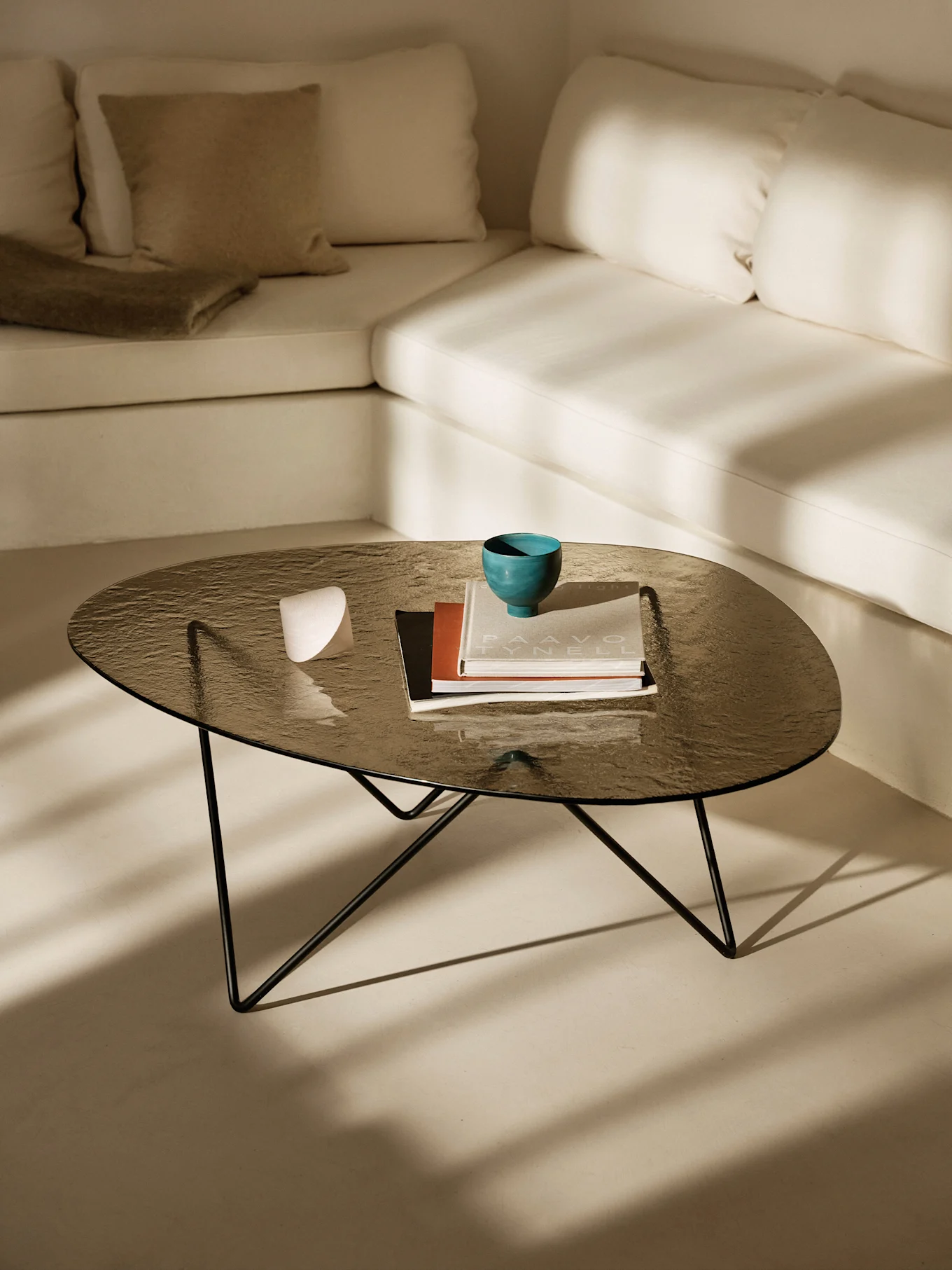 Mørkt glas sofabord med en turkis skål, bøger og et hvidt objekt. Lys L-formet sofa og sollys skygger.