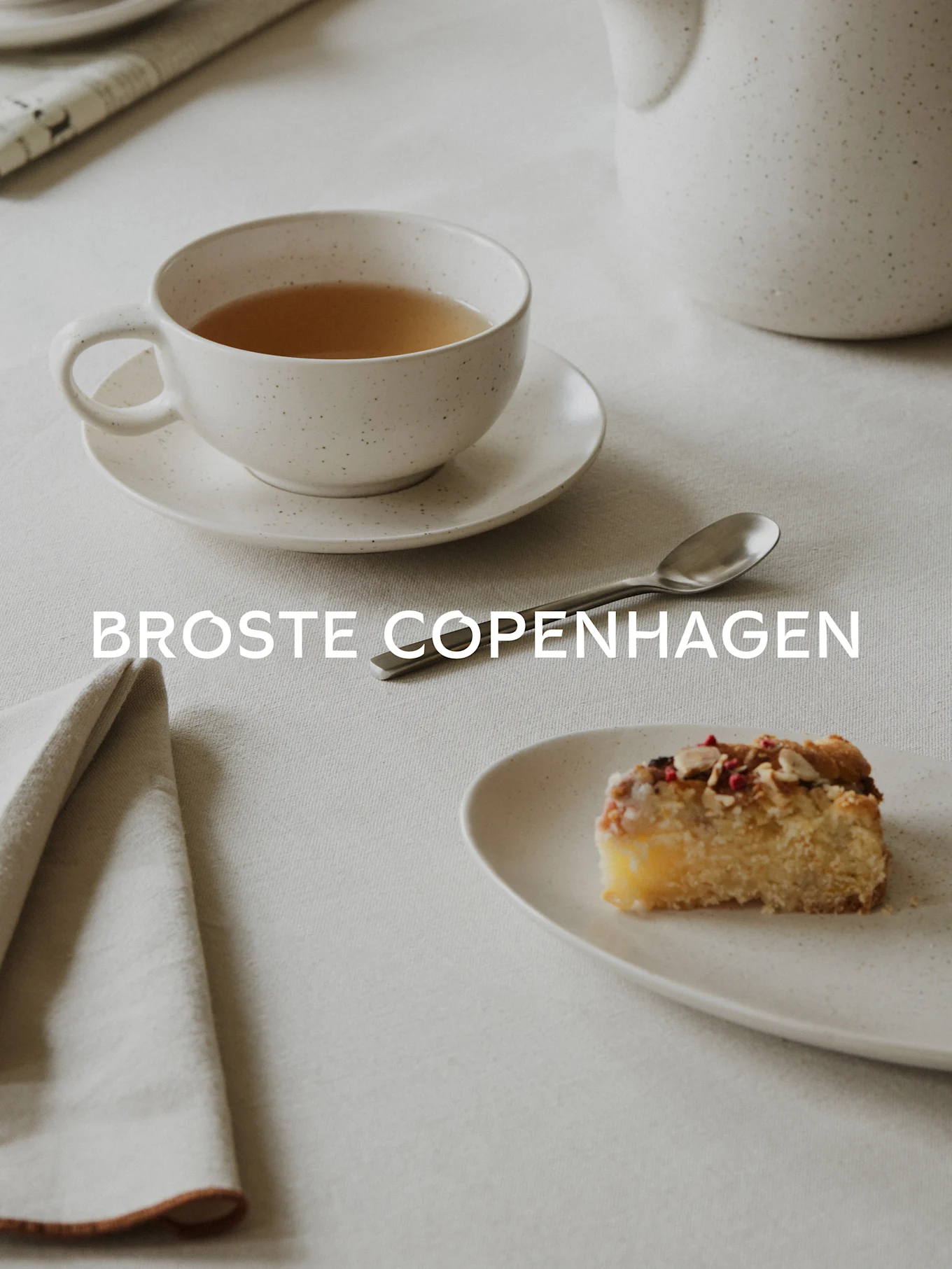 Te i en plettet hvid kop, et stykke kage med mandler og bær på en matchende tallerken, en ske og en serviet på en linneddug. Tekst: BROSTE COPENHAGEN.