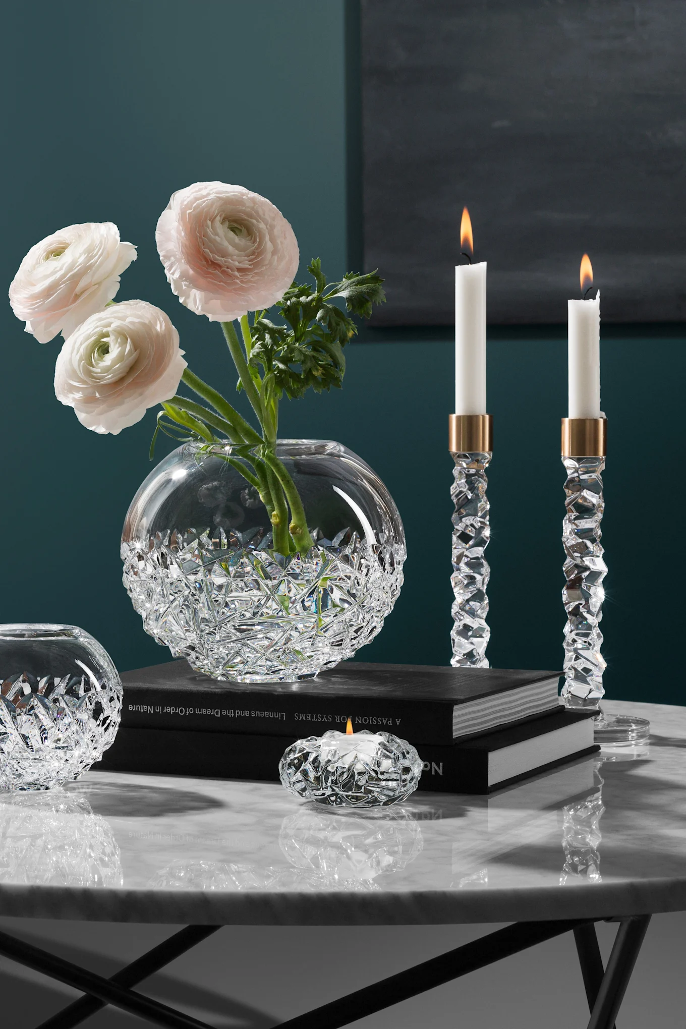 Elegant krystalvase med lyserøde ranunkler, to tændte krystalstager og en fyrfadsstage på et marmorbord.