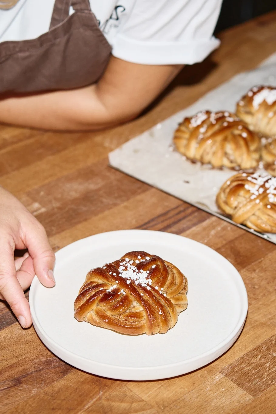 Hånd lægger en sukkerstrøet, flettet kanelsnegl på en tallerken; flere snegle på en bakke.