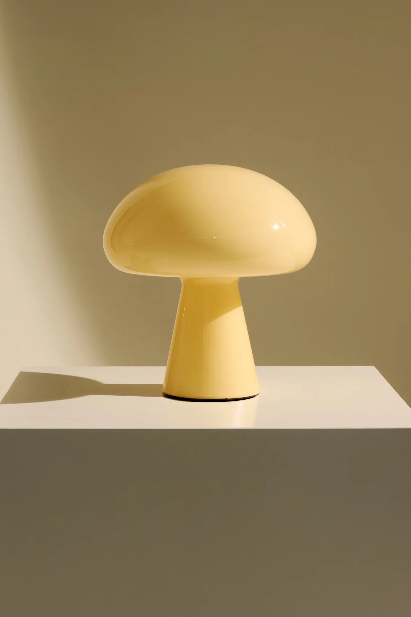 En lysegul champignonformet lampe på en hvid sokkel mod en lysebrun baggrund.