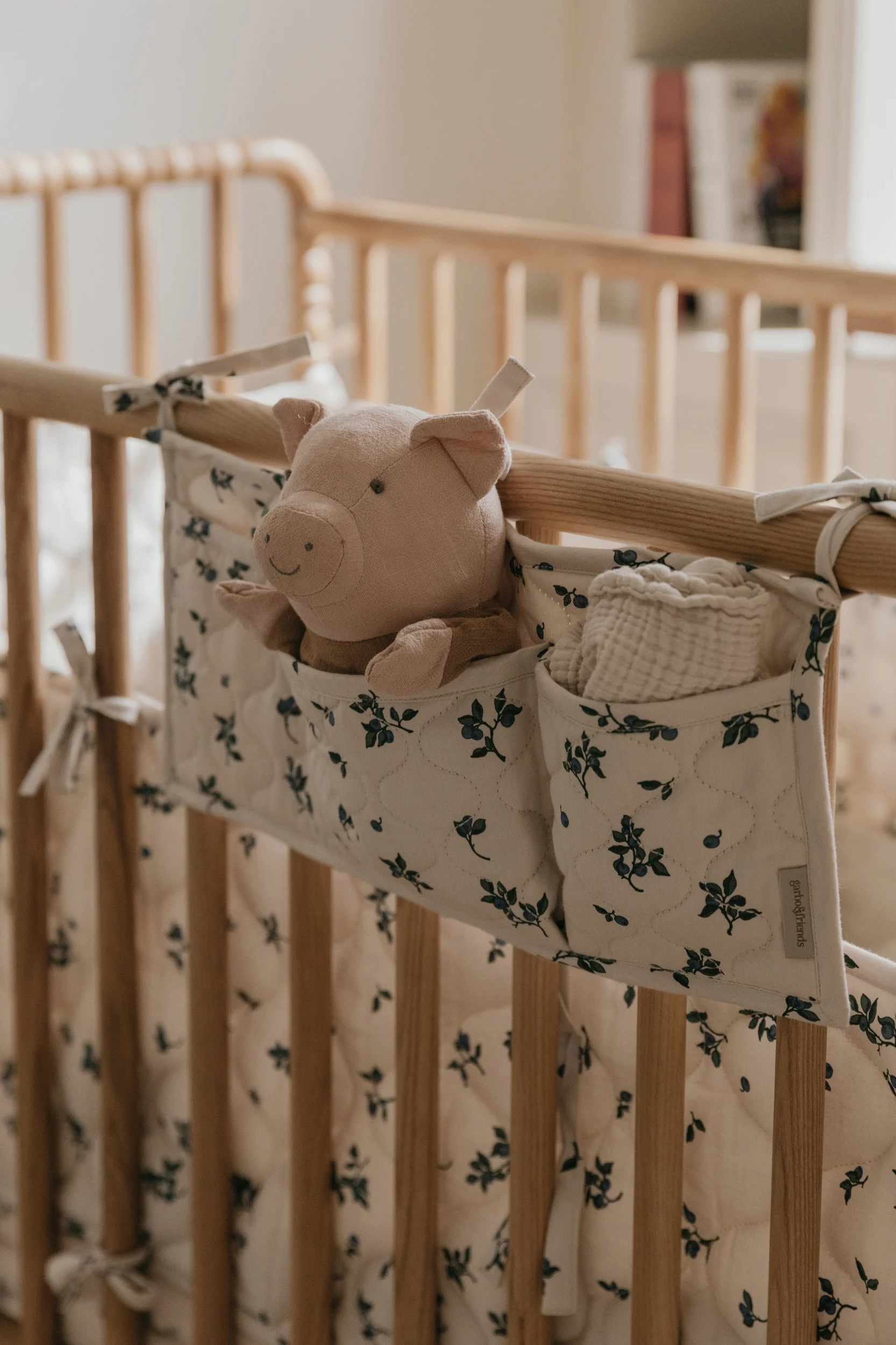 En lyserød plysgris og en hvid muslin klud i en cremefarvet og blå blomstret organizer hængende på en tremmeseng af træ.