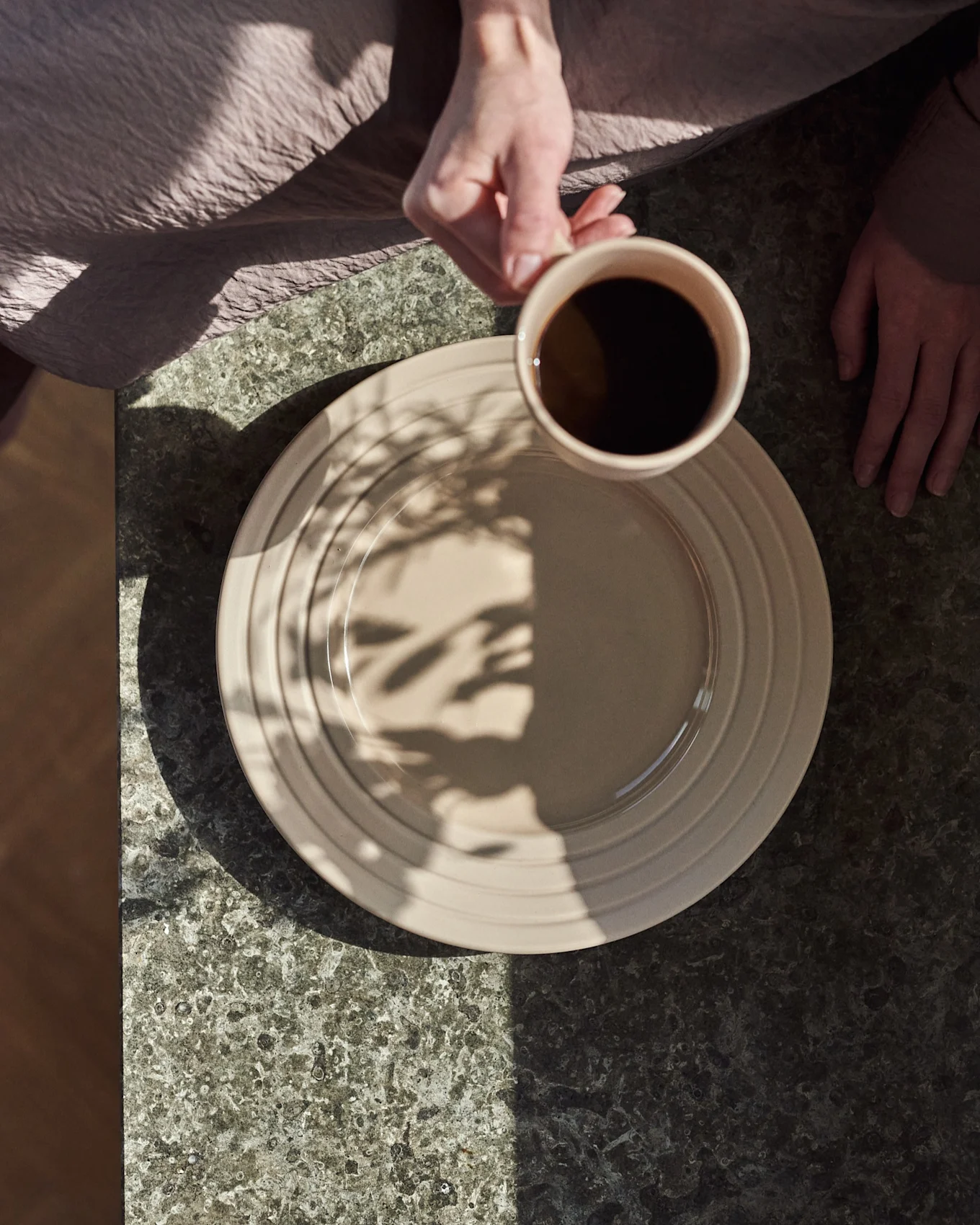 Ovenfra: hænder, en holder et beige krus med sort kaffe over en beige tallerken på et mørkt stenbord med bladskygger.