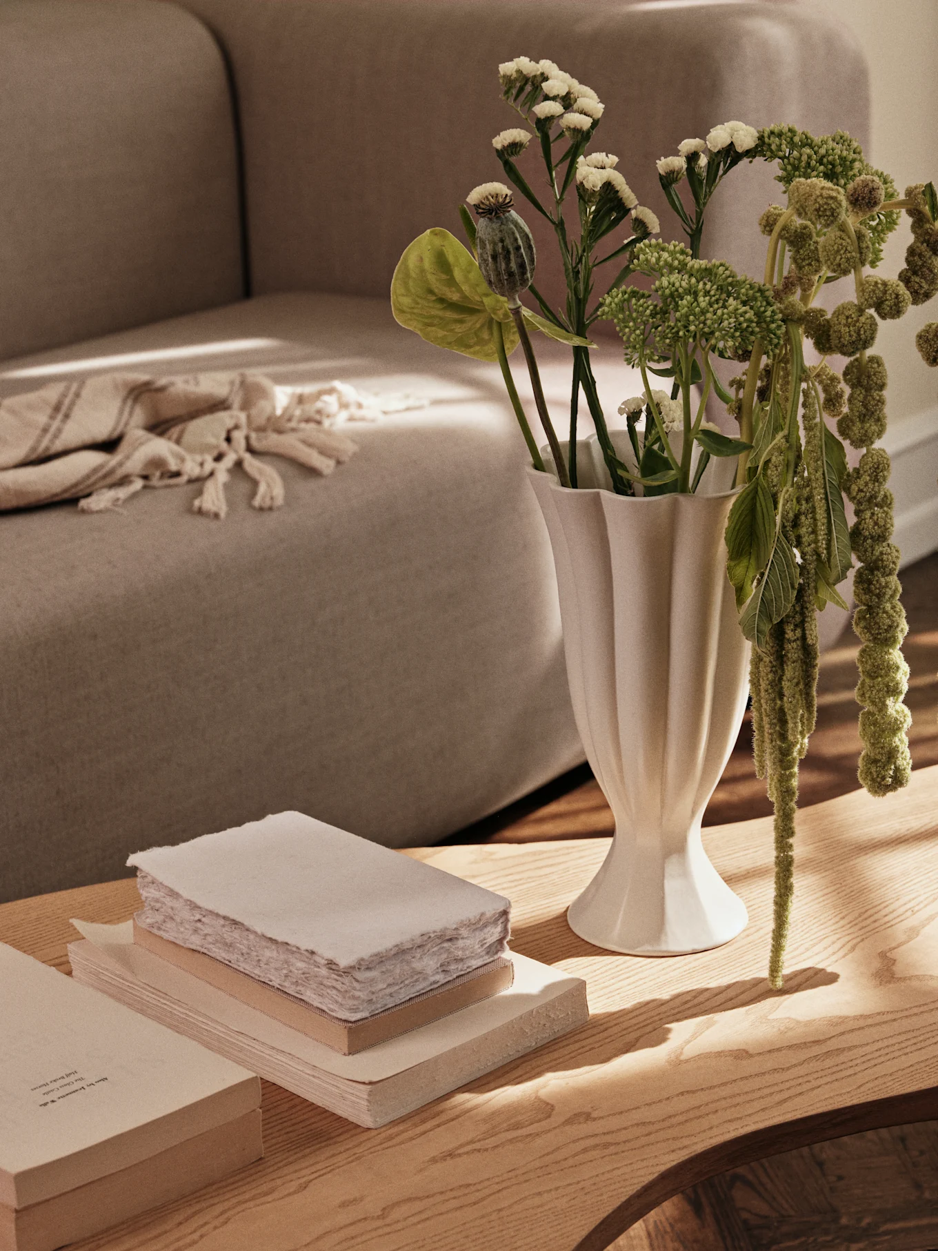 En hvid riflet vase med grønne planter og en stak bøger på et sofabord i træ, med en sofa i baggrunden.