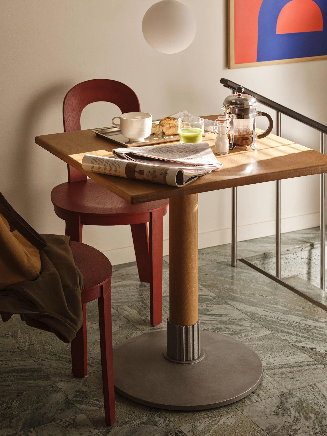 Et lille træspisebord med morgenmad, inklusive en kaffekande, en kop, en croissant, en grøn drink og aviser. To røde stole står ved bordet, og trapper med gelænder er i baggrunden.