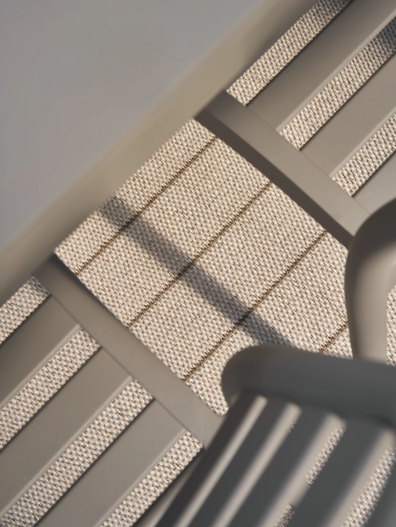 Nærbillede, vinklet udsigt over lyst beige vævet materiale med diskrete vandrette og diagonale brune striber, indrammet af glatte grå bjælker.