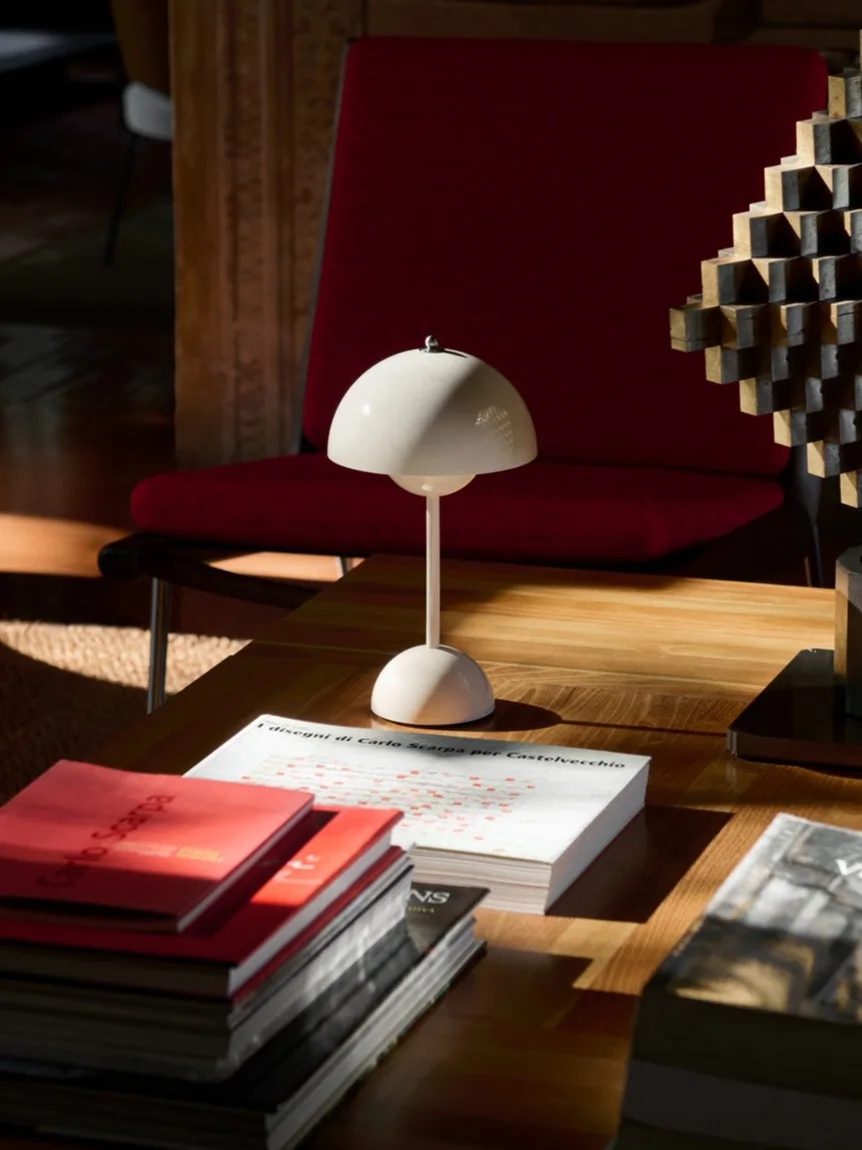 En hvid svampeformet bordlampe, bøger og en geometrisk skulptur på et sofabord af træ, med en rød lænestol bagved.