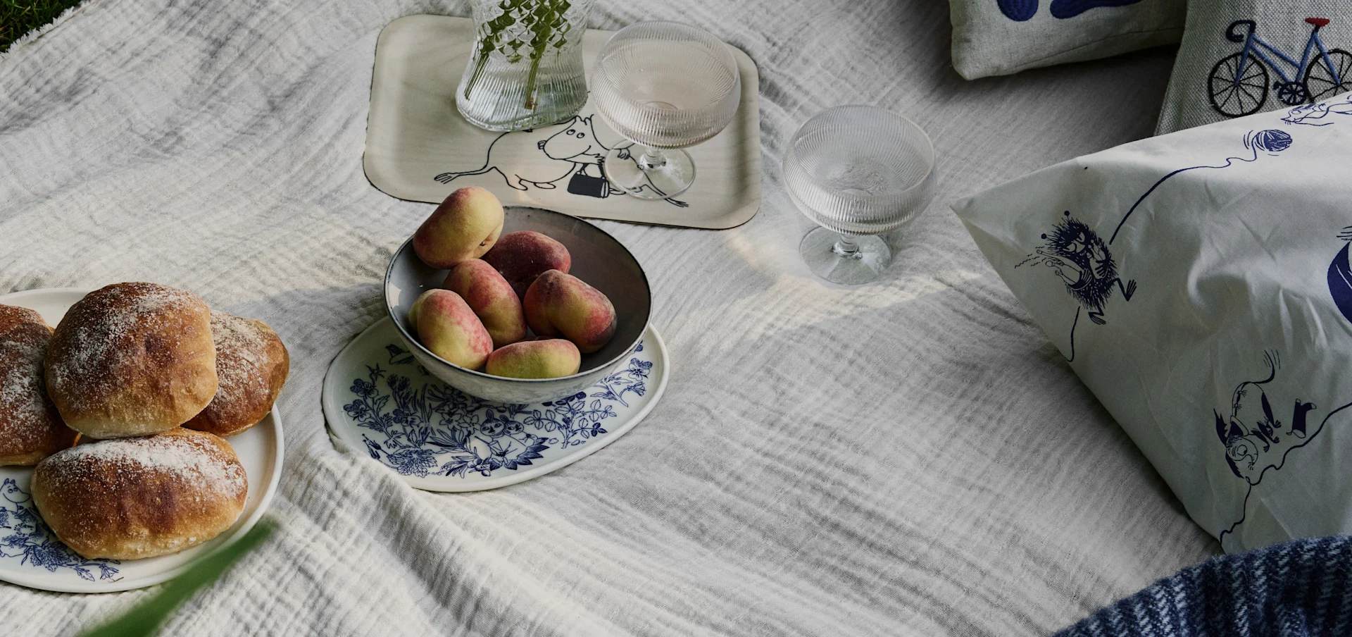 Picnicscene på et hvidt tæppe med brødboller, ferskner, drikkevarer og puder med Moomin- og cykelmønstre.