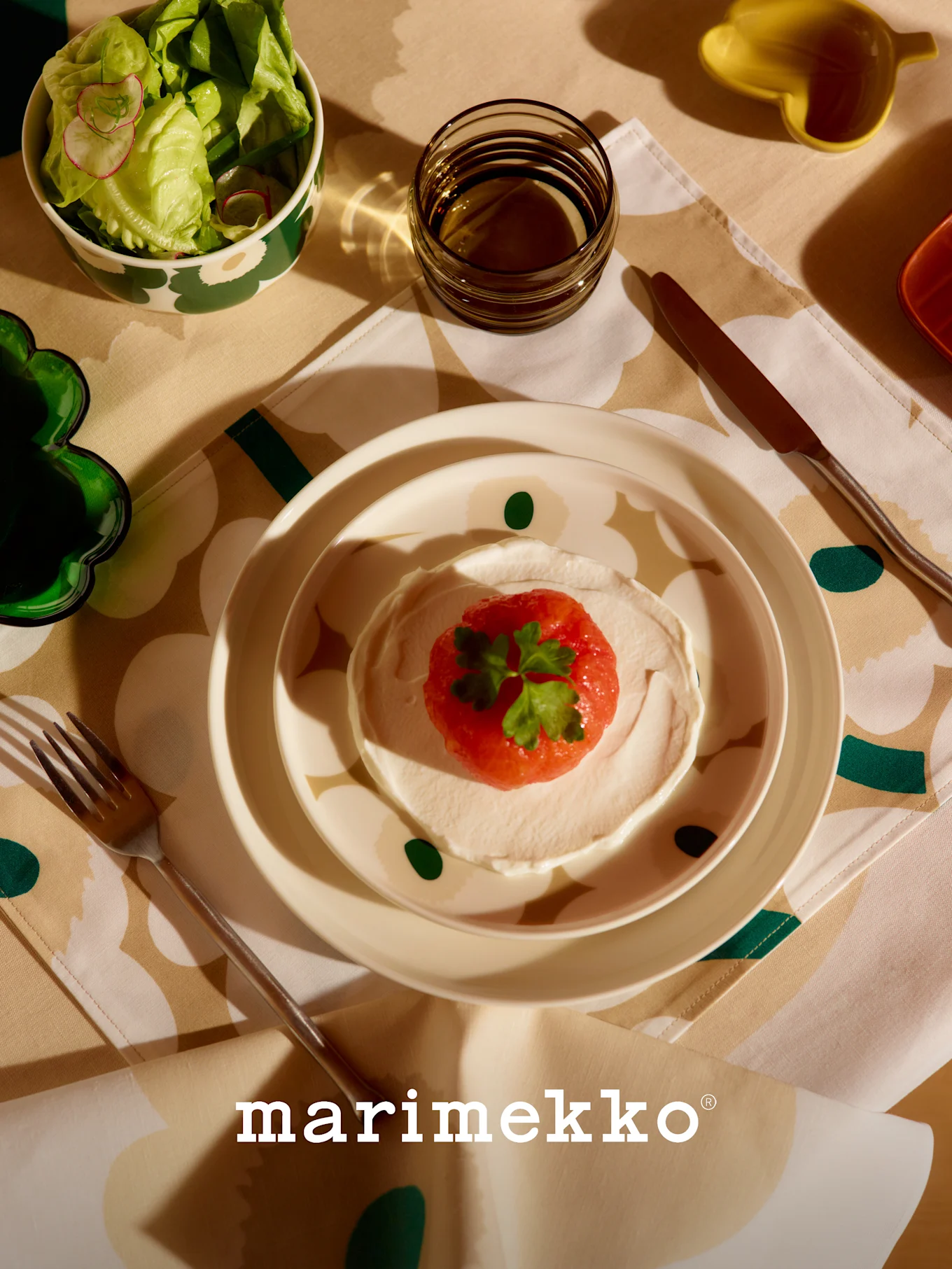 Marimekko borddækning med mønstret salatskål, ret med rød frugt og creme, glas og bestik på en blomstret dug.