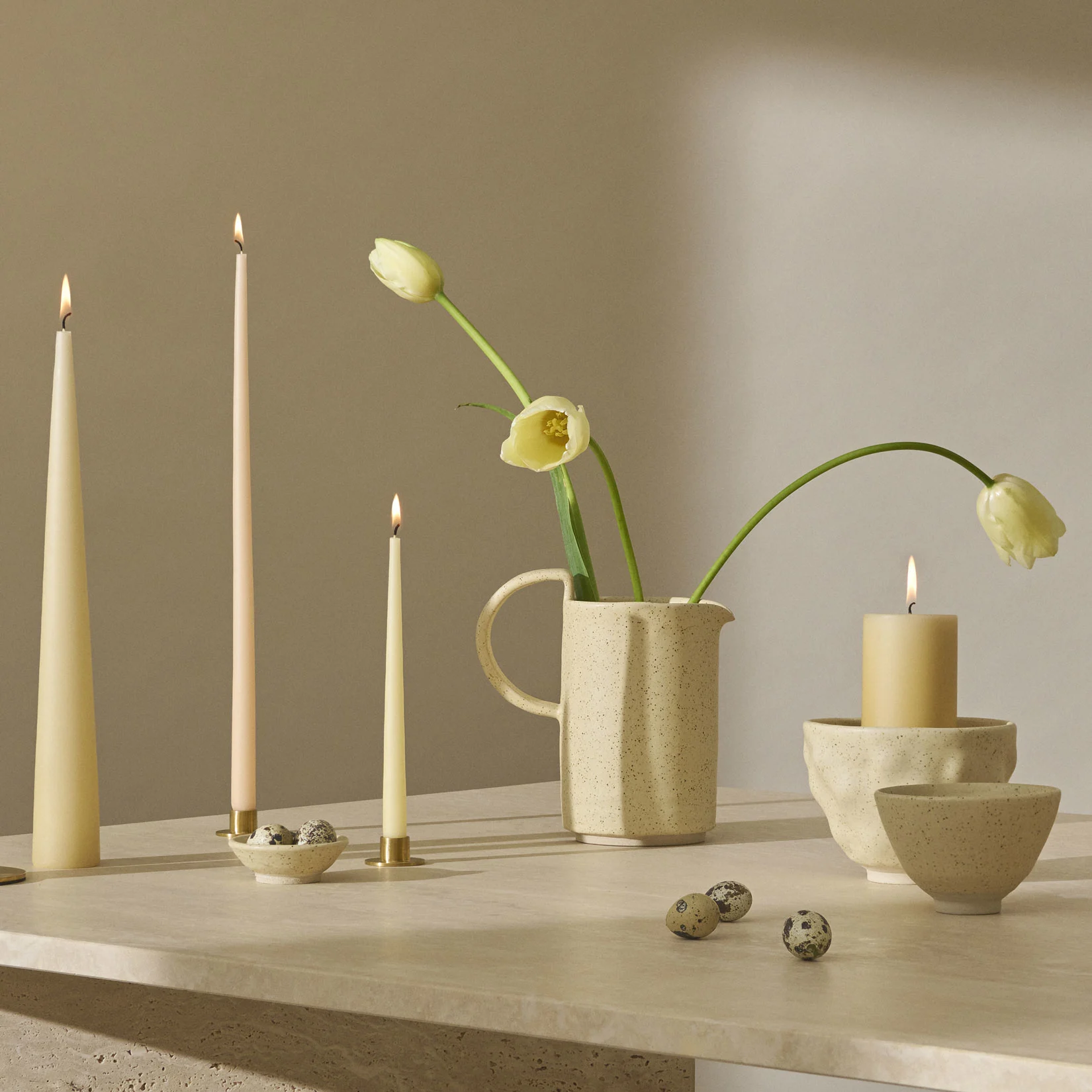 Et stilleben med tændte stearinlys, lysegule tulipaner i en plettet kande og vagtelæg på en tekstureret overflade.