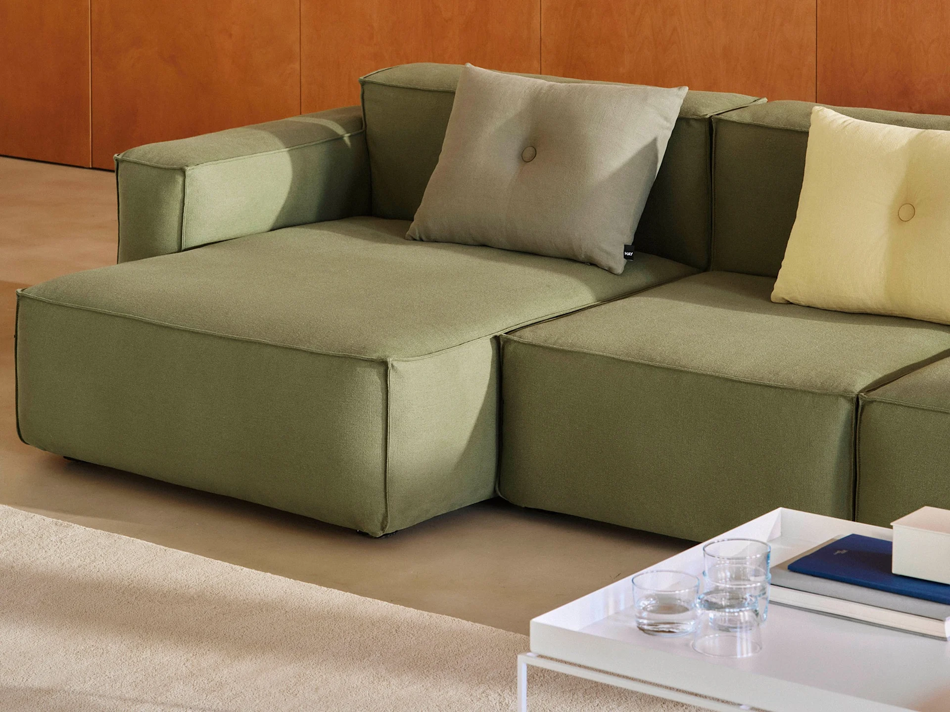 En modulær olivengrøn sofa med lysegrå og gule pyntepuder, ved siden af et hvidt sofabord med bøger.