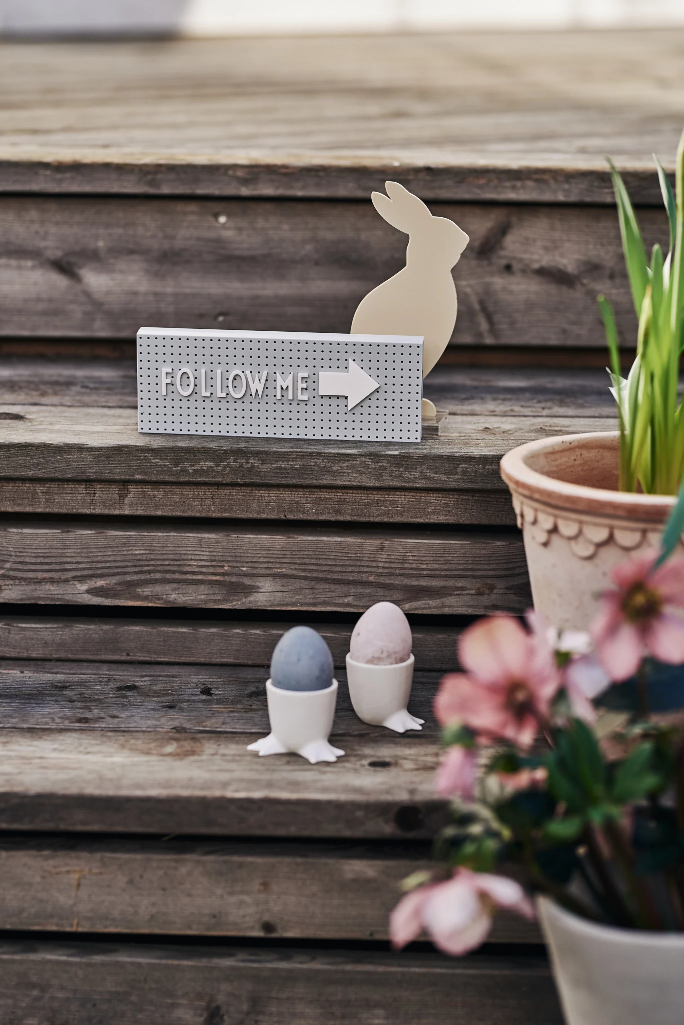 Naturligt farvede påskeæg står i to hvide vandrende æggebægre fra DBKD på en veranda, med blomster i potter ved siden af og The Sign-bogstavtavlen fra Design Letters bag dem.
