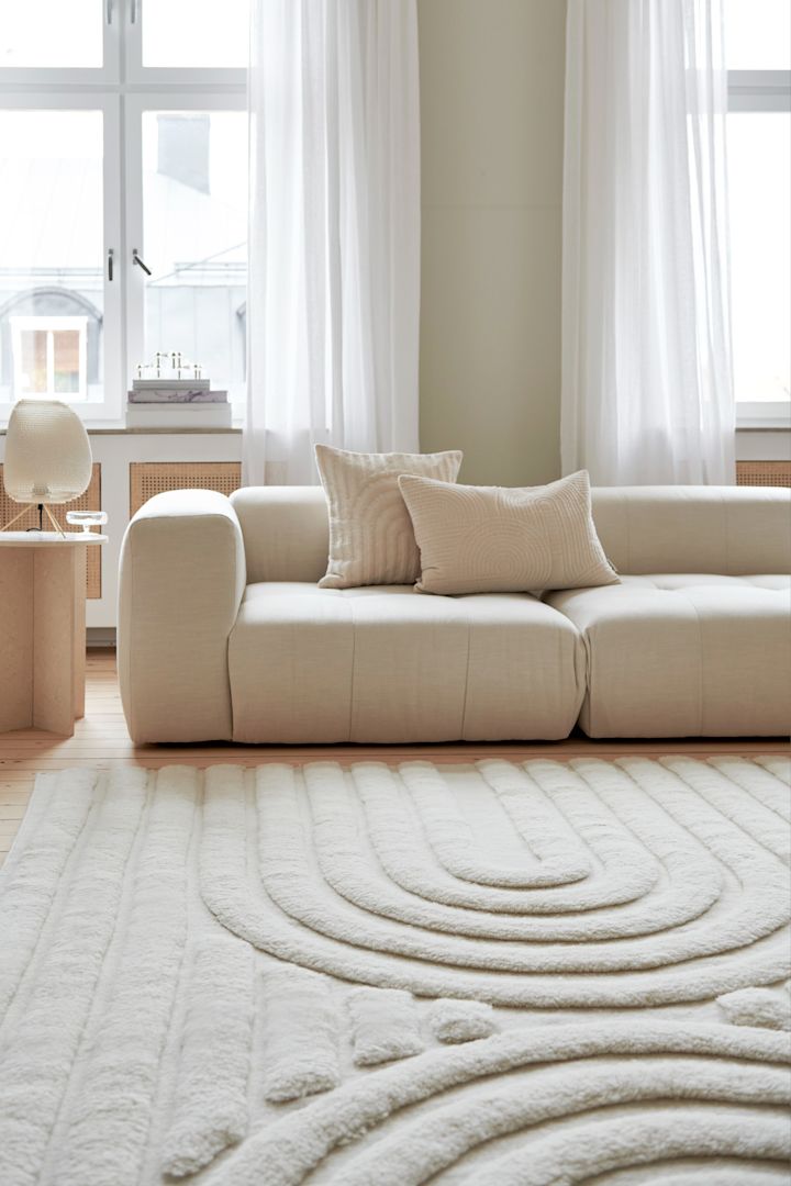 Tæpper til stuen: Det lyse Curve-uldtæppe fra Classic Collection ligger foran en sofa i en lys stue.