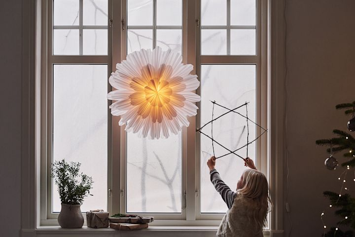 Snöblomman adventsstjernen fra Watt & Veke i et vindue, et barn står ved siden af og holder en stjerne lavet af pinde.