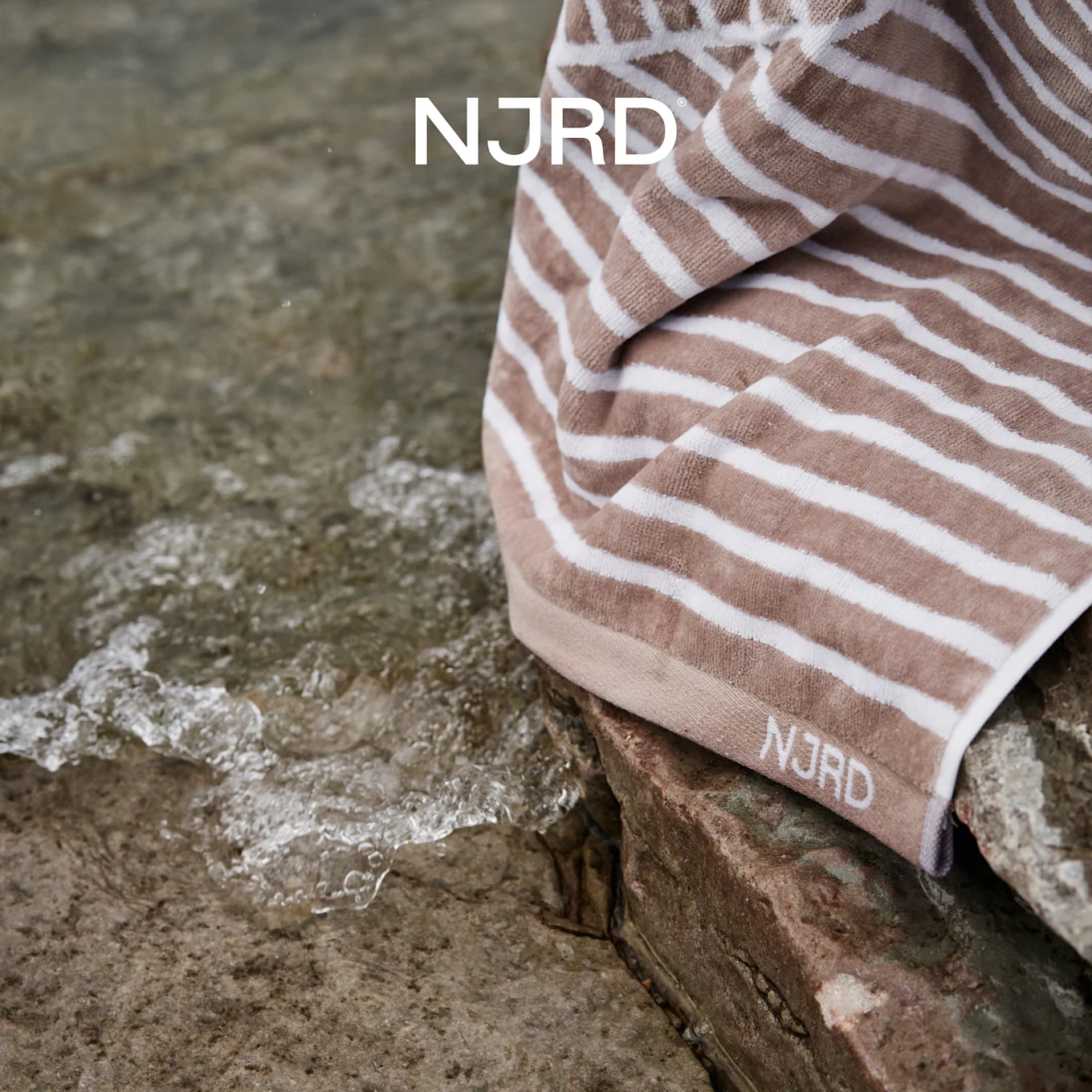 NJRD beige og hvidstribet håndklæde på klipper ved krusende vand.
