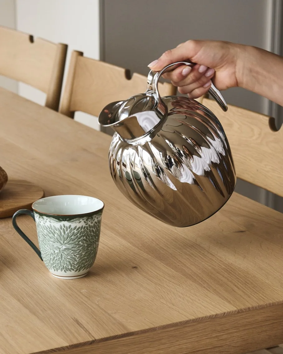 En hånd hælder fra en sølvfarvet riflet kande ned i et hvidt krus med grønt bladmønster på et træbord.