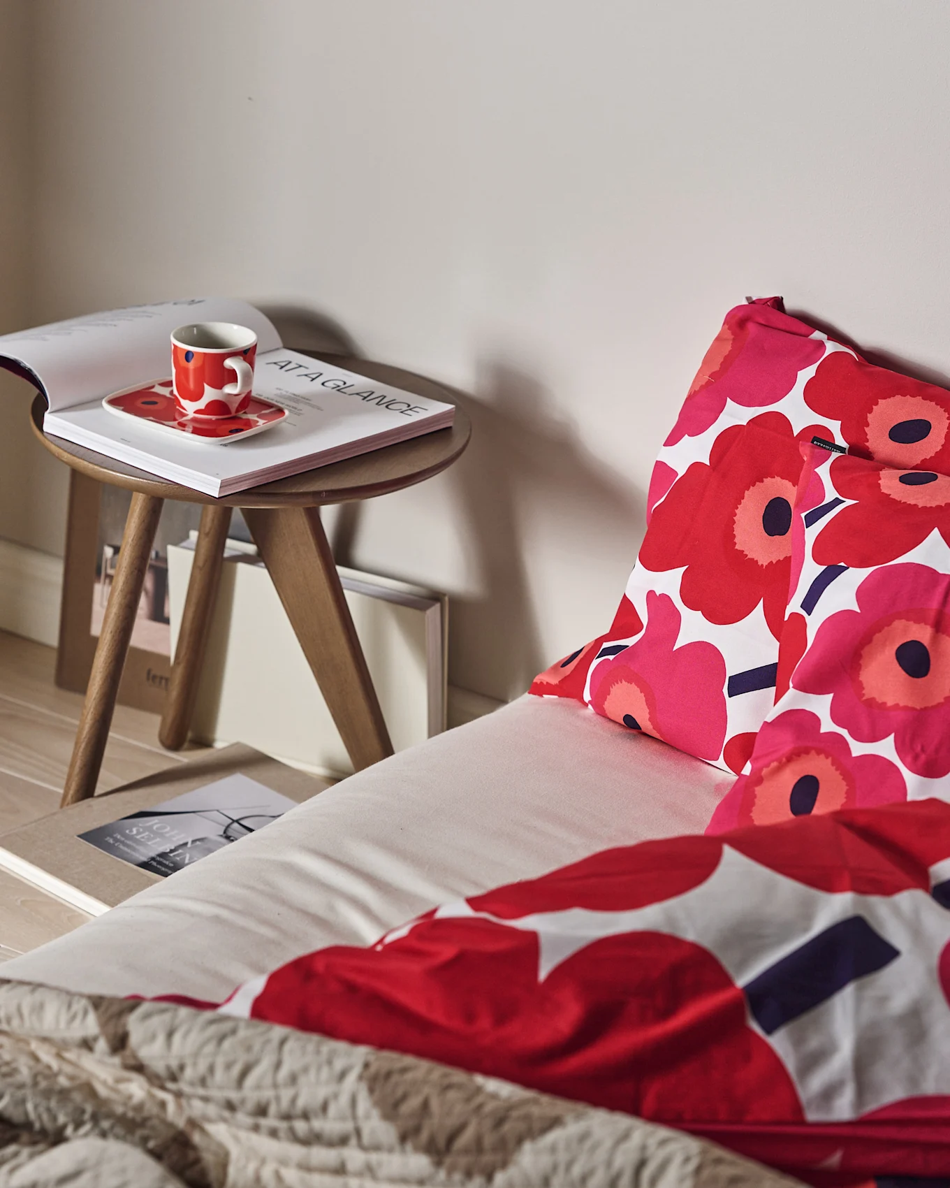 En seng med rød og pink blomstret sengetøj og et tekstureret beige tæppe. En rød kop og åben bog på et træsidebord.