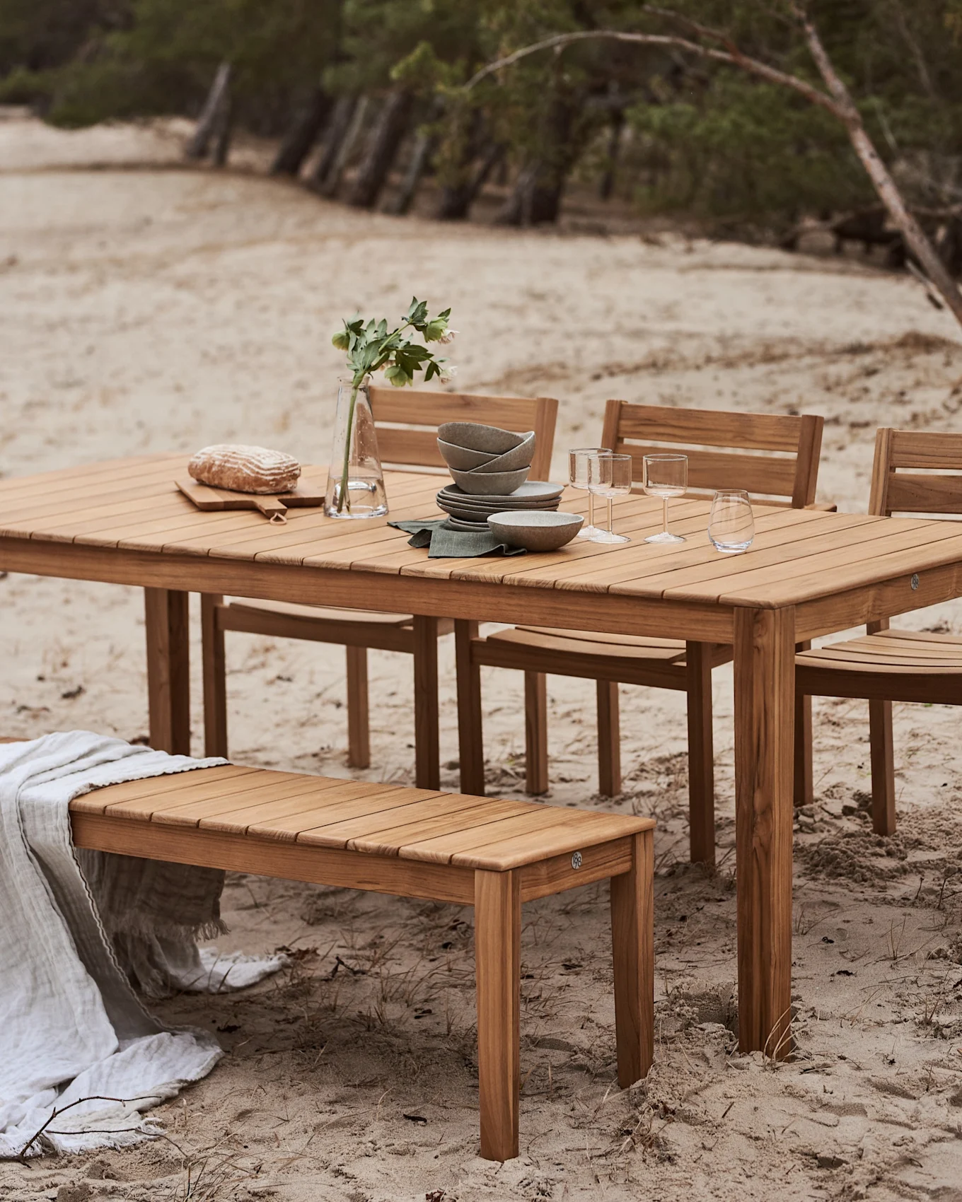 Udendørs spisesæt i træ med bord og bænk på en sandstrand. Bordet er dækket med service, glas, brød og en vase med blomster.