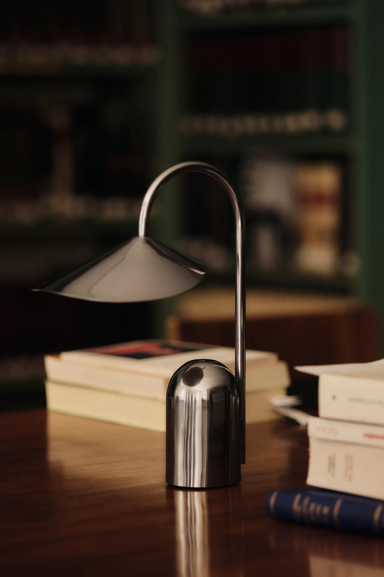 En moderne sølv bordlampe med buet stilk og konisk skærm står på et træskrivebord med slørede bøger i baggrunden.