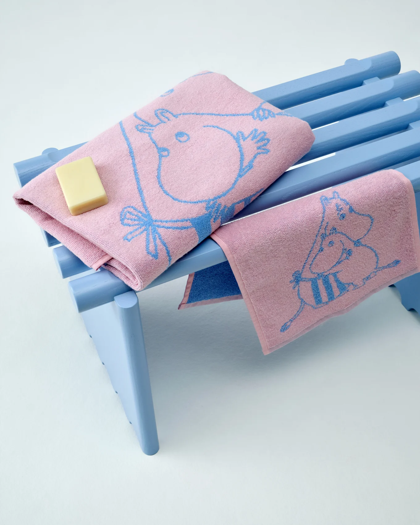 To lyserøde Moomin-håndklæder med blå mønstre, det ene foldet med et stykke sæbe, på en lyseblå bænk.