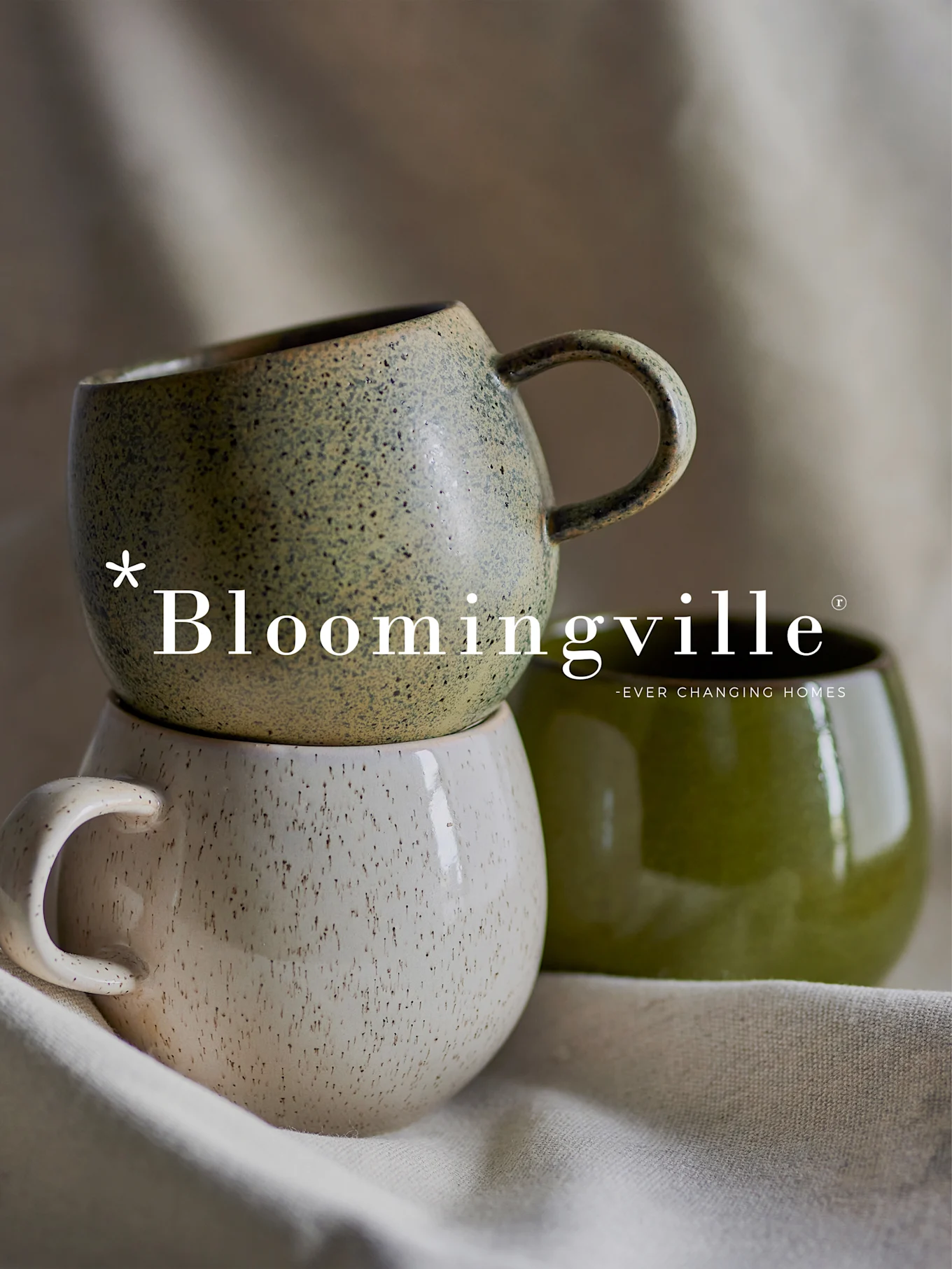 Bloomingville logo over en stabel af to plettede keramikkrus i grøn og råhvid, med et andet ensfarvet grønt krus ved siden af.