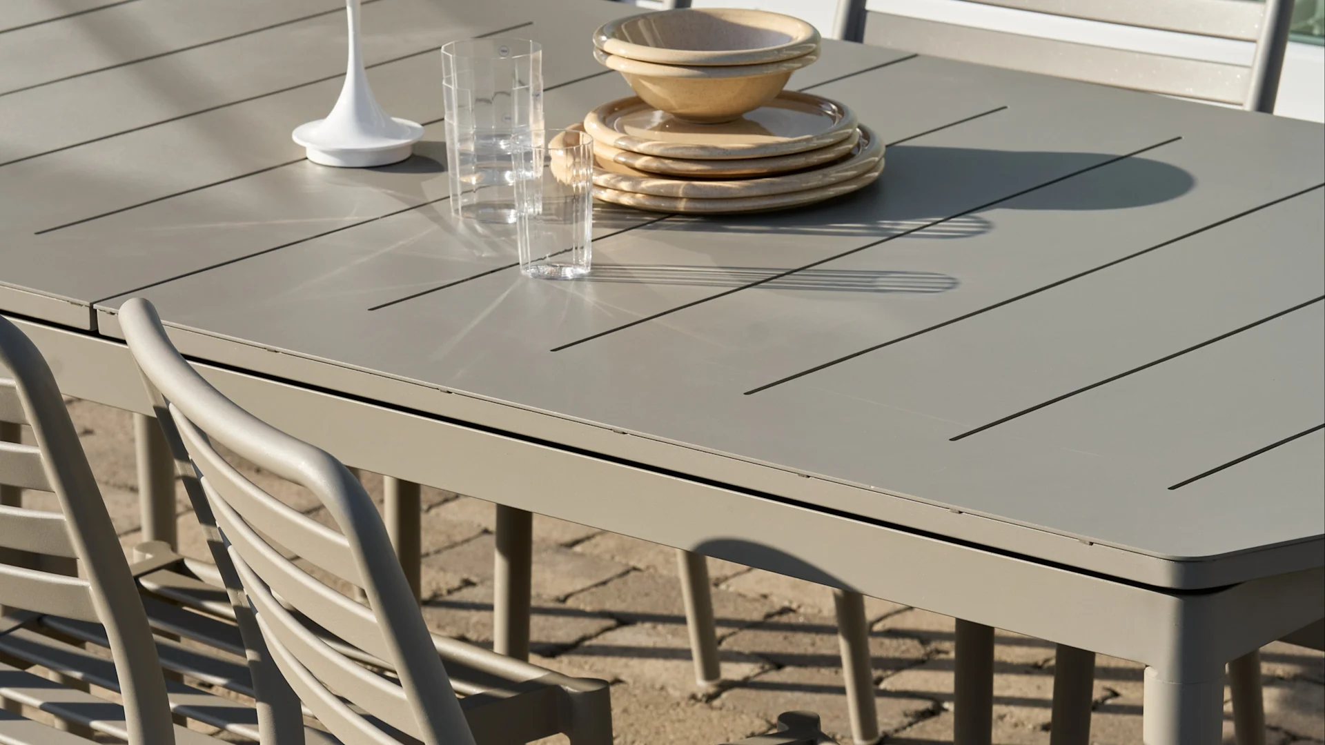 Lyst gråt lamel udendørs spisebord med stablede beige tallerkener og skåle, to klare glas og en hvid pyntegenstand.