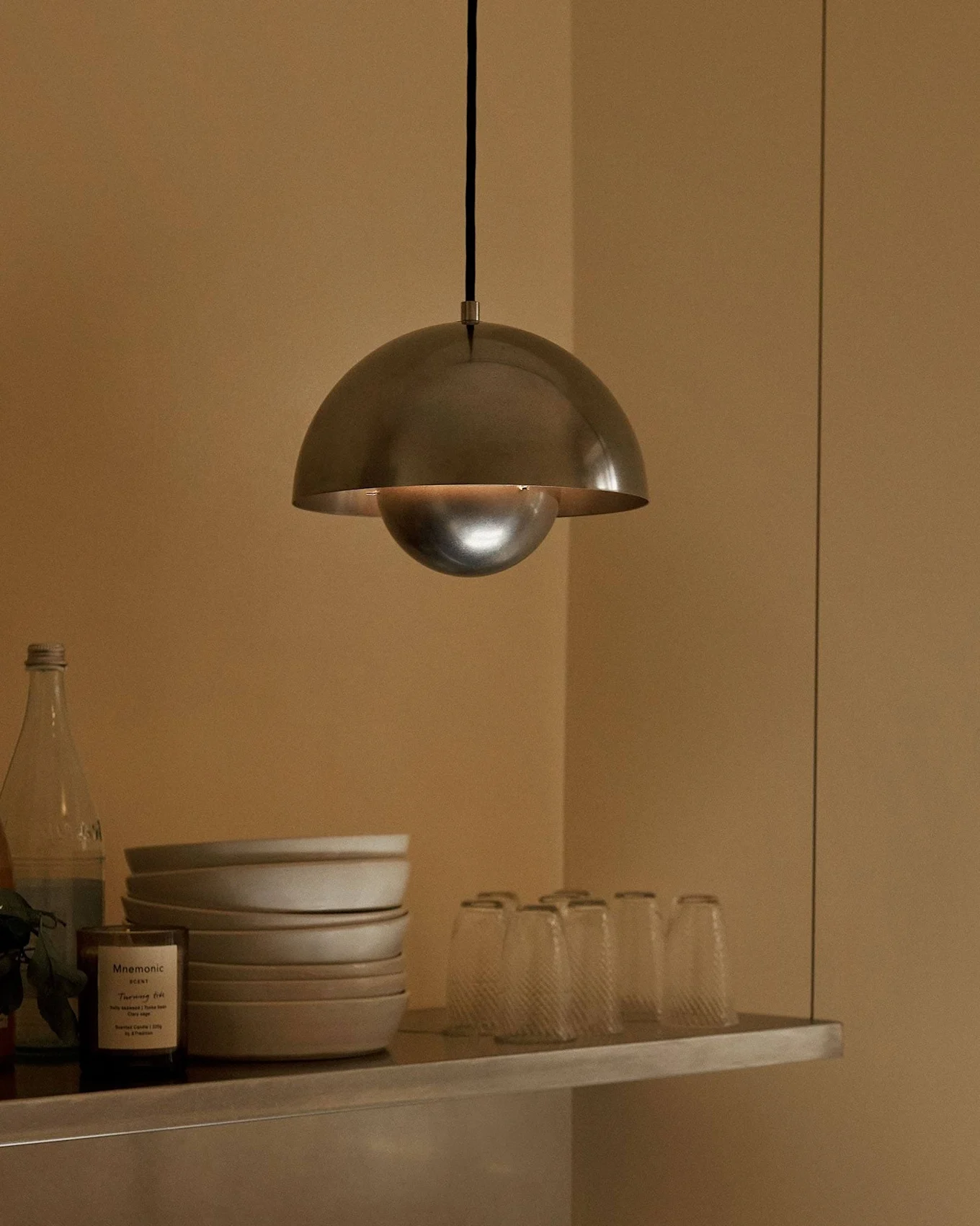En kuppelformet pendellampe i metal hænger over en hylde med skåle, glas, flasker, en frugt og et duftlys.