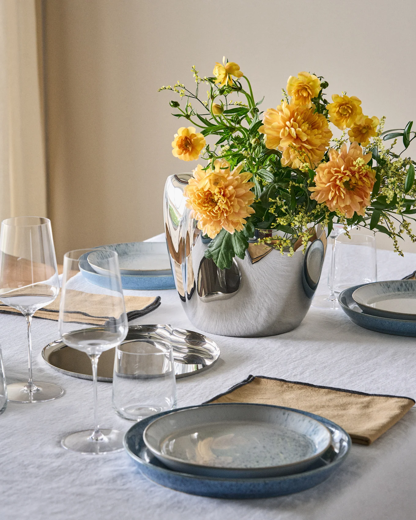 Et spisebord dækket med blå tallerkener, beige servietter, klare glas og en sølvvase med orange-gule blomster på en hvid dug.