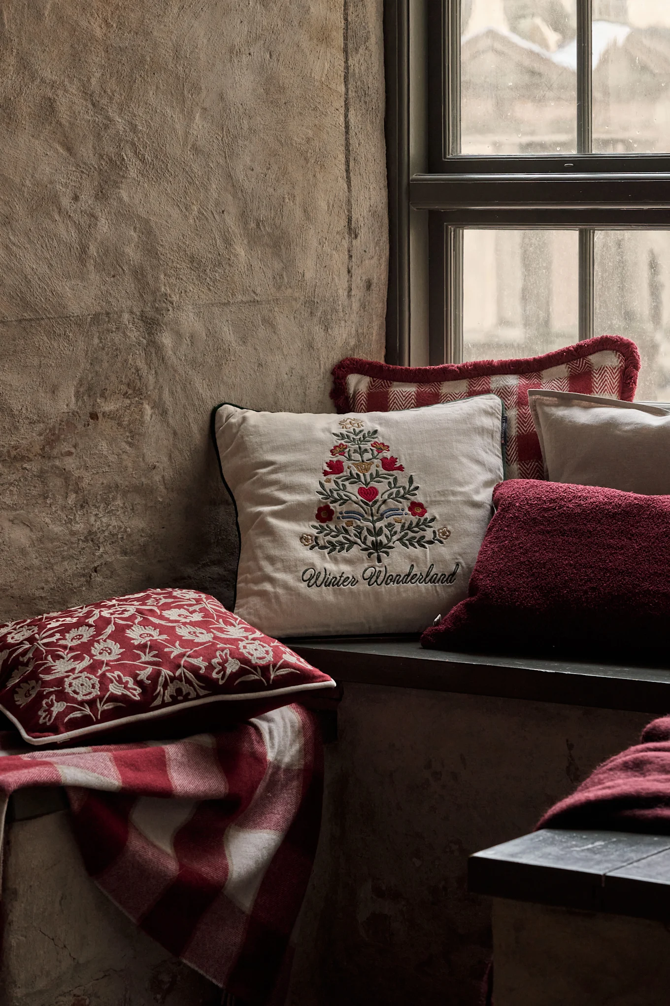 Hyggelig vindueskrog med festlige puder, inklusive en med et broderet juletræ og 'Winter Wonderland', og et rødt ternet tæppe.