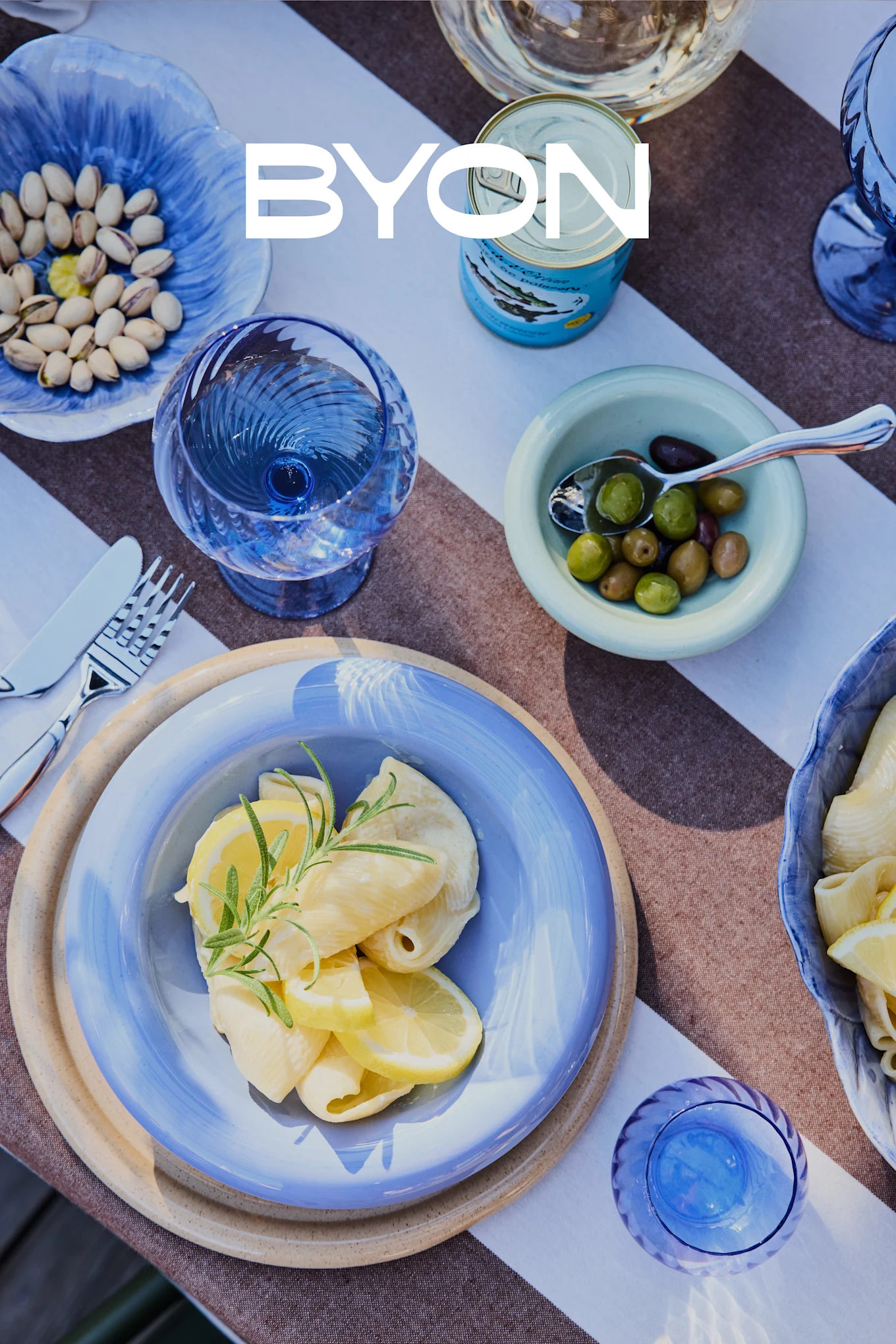 Oversigt over et spisebord med blåt service, inklusive en tallerken pasta med citron og rosmarin, skåle med pistacienødder og oliven samt blå glas. BYON-logoet er overlejret.