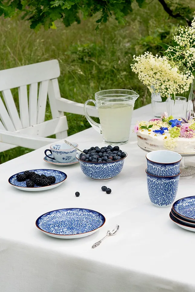 Udendørs borddækning med blå og hvide mønstrede retter, friske blåbær og brombær, en limonadekande og en blomsterdekoreret kage.
