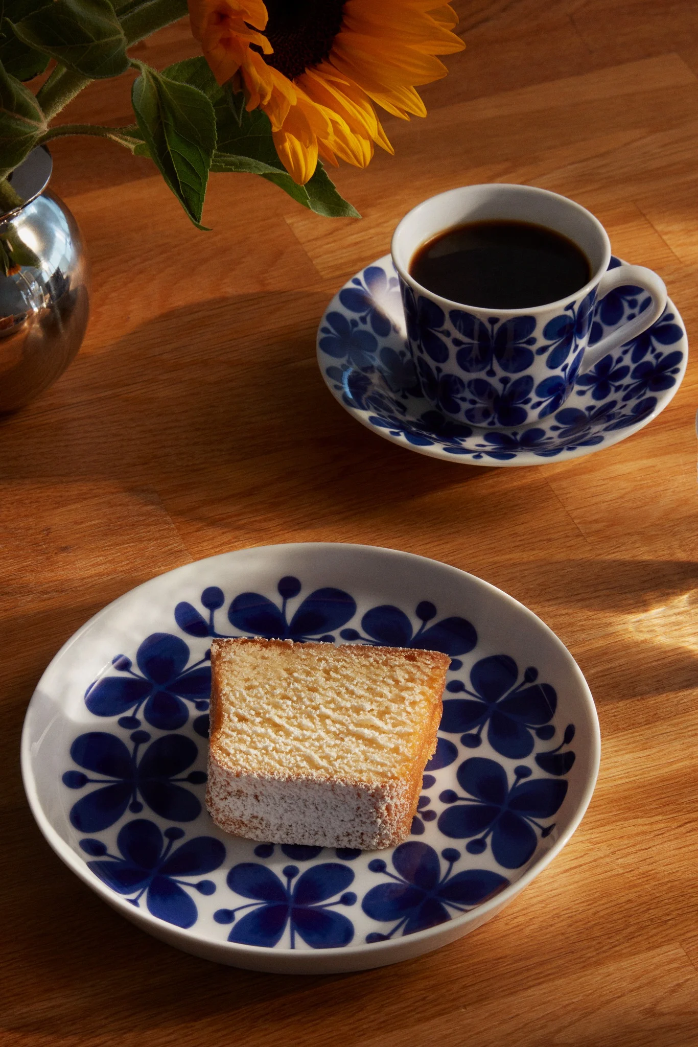 Pudderkage på en blå blomsterplade, sort kaffe i en matchende kop og en solsikke i en sølvvase på træ.