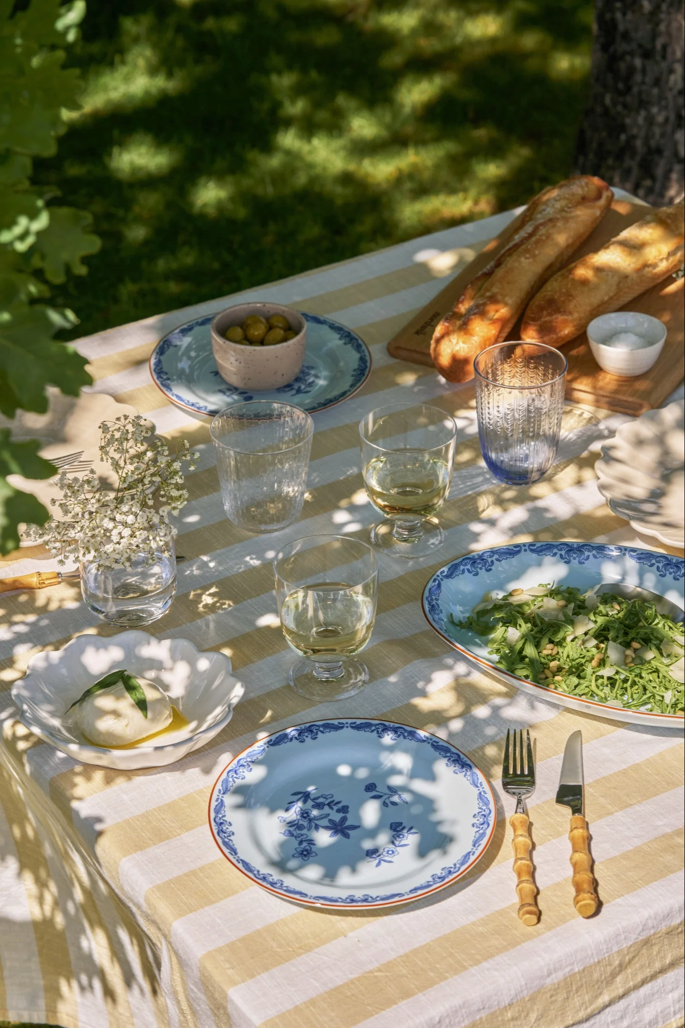 Et udendørs picnicbord dækket med en gul og hvid stribet dug, burrata, salat, baguetter, oliven og blåmønstrede tallerkener.