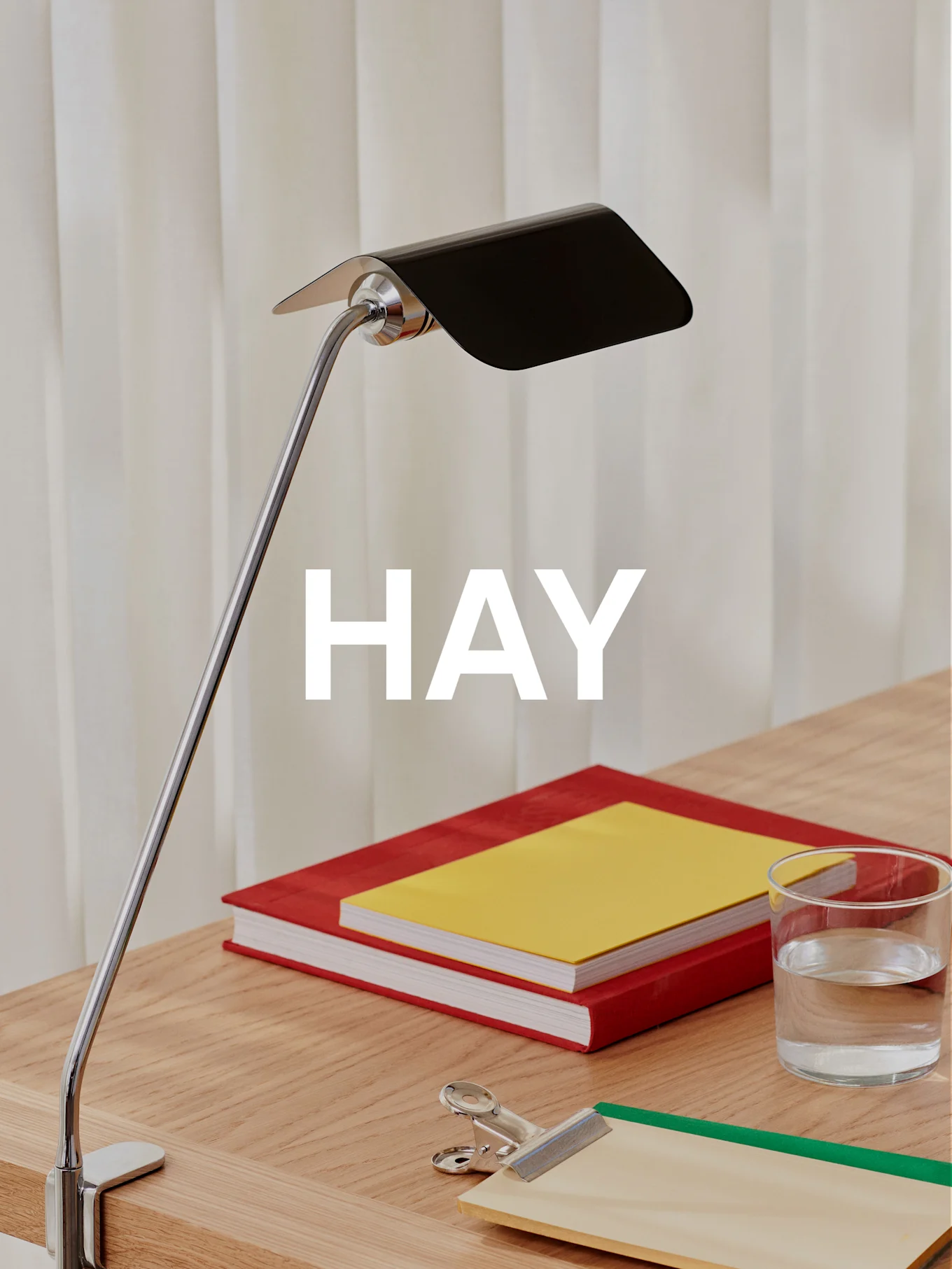 En krom bordlampe med sort skærm belyser et træskrivebord med to bøger, et glas vand og et clipboard.
