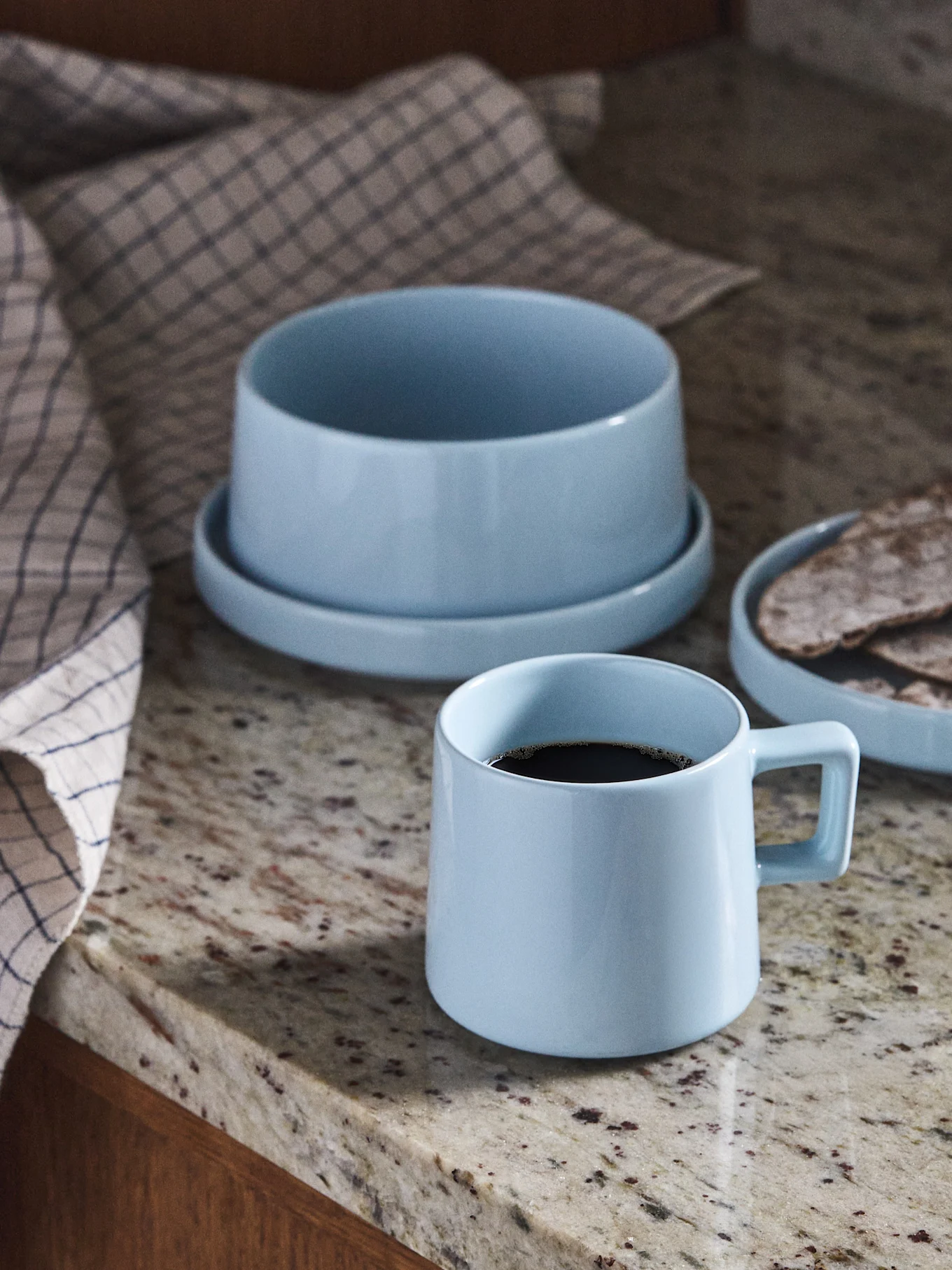Lyseblå keramisk kaffekop, skål og tallerken med fladbrød på en granitbordplade.