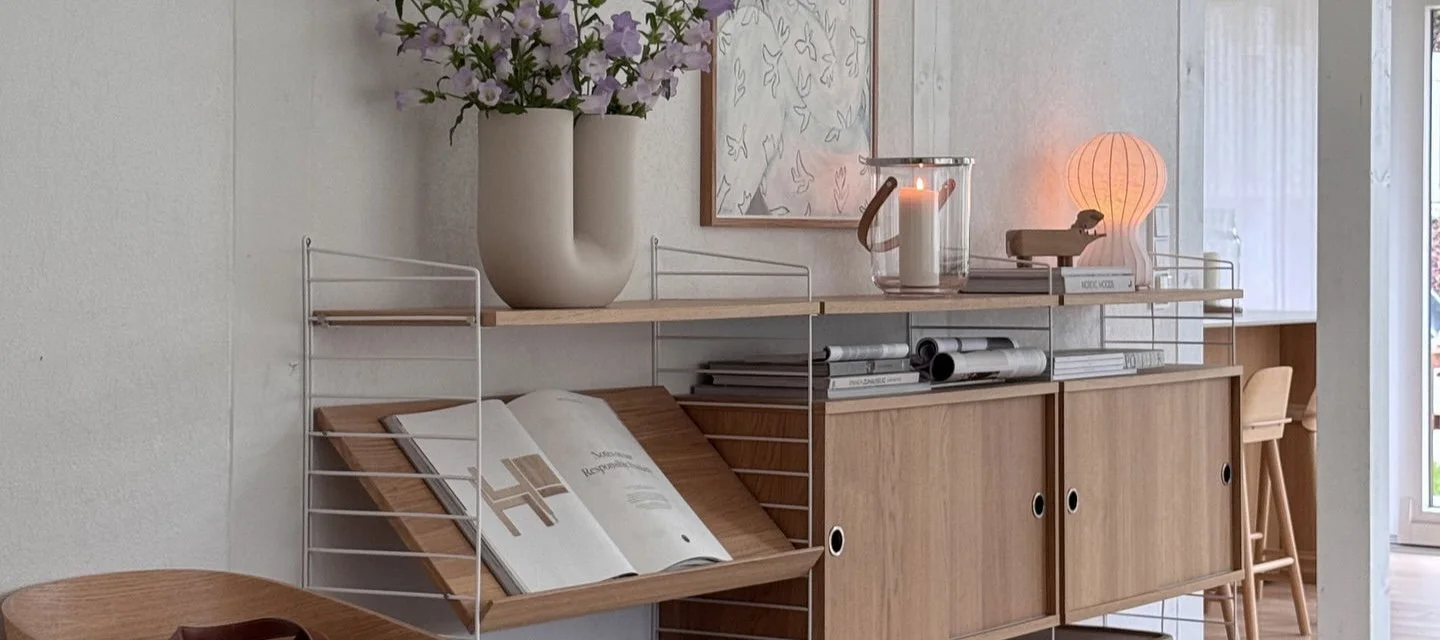 Modulært reolsystem med hvide rammer og lyse træhylder, skabe, vase med lilla blomster, tændt lanterne og glødende lampe.