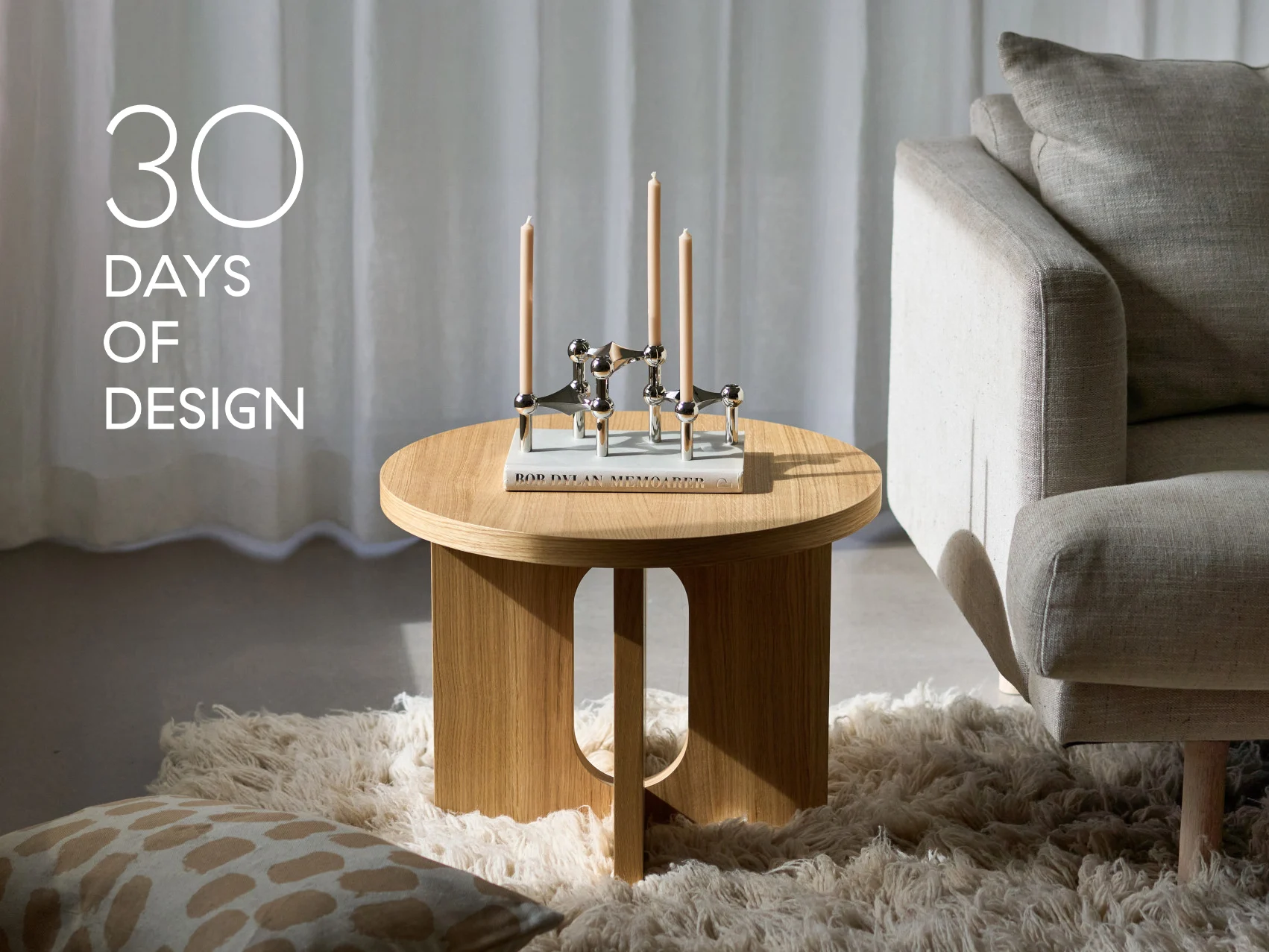 Moderne stue med rundt træsidebord, sølvlysestage med tre stearinlys, en bog og en beige sofa. Tekst: '30 DAGE MED DESIGN'.