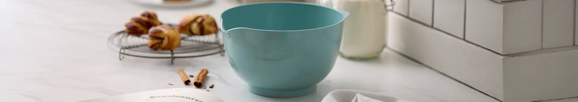 Lyseblå røreskål, bagværk på rist, mælkeflaske og opskriftsbog på marmorbordplade.