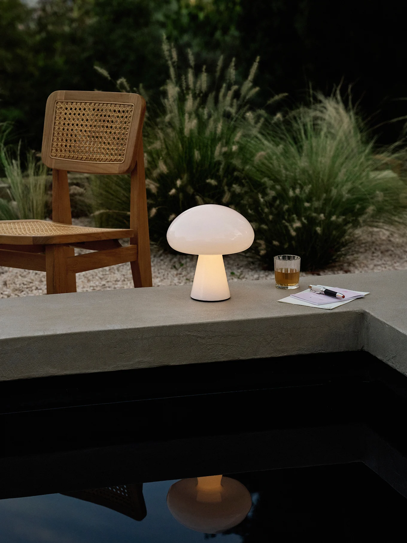 En glødende champignonformet lampe på en betonkant ved siden af en pool, med en træstol, drik og kuglepen.